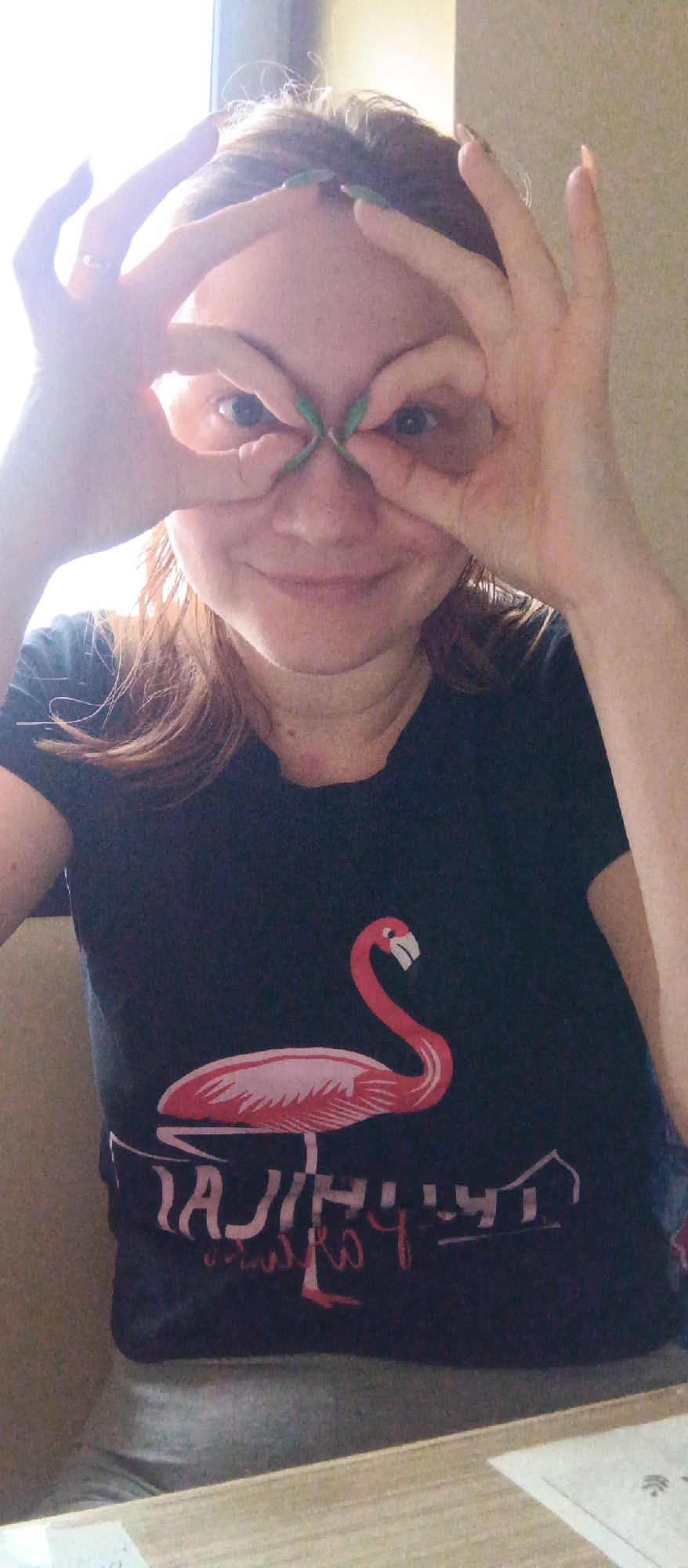 A medium-shot photograph captures a fair-skinned woman indoors, possibly in Unknown, Unknown, wearing a dark t-shirt adorned with a pink flamingo graphic and an illegible, backwards white inscription beneath it. The woman is positioned against a neutral background. Her brown hair is casually styled, and she is looking directly into the camera with a playful expression. She has shaped her fingers into circles, placing them over her eyes like makeshift spectacles. Her long fingernails are adorned with green polish. The environment includes a light-colored wooden table in the foreground and glimpses of natural light filtering through a window.