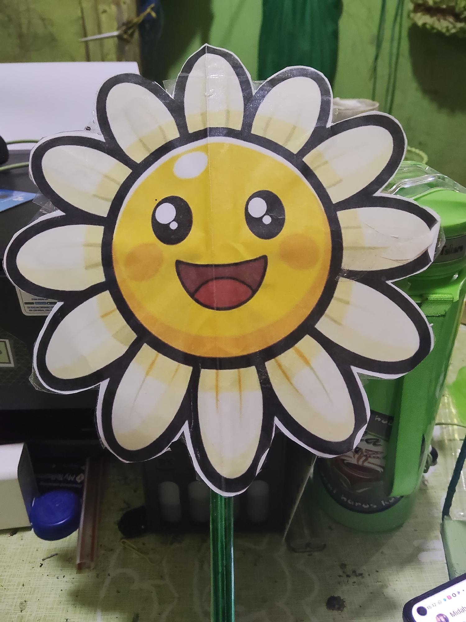 A close-up, eye-level shot shows a handmade, cartoon-like sunflower decoration. The sunflower has a yellow face with large, black eyes, rosy cheeks, and an open, smiling mouth with a red tongue. It has white petals outlined in black, with subtle yellow shading. The decoration is mounted on a green stick. The background is blurred, but appears to be an indoor setting with a green wall or fabric, and various objects including a white box and a green container. There are no people present in the image. The lighting is artificial.