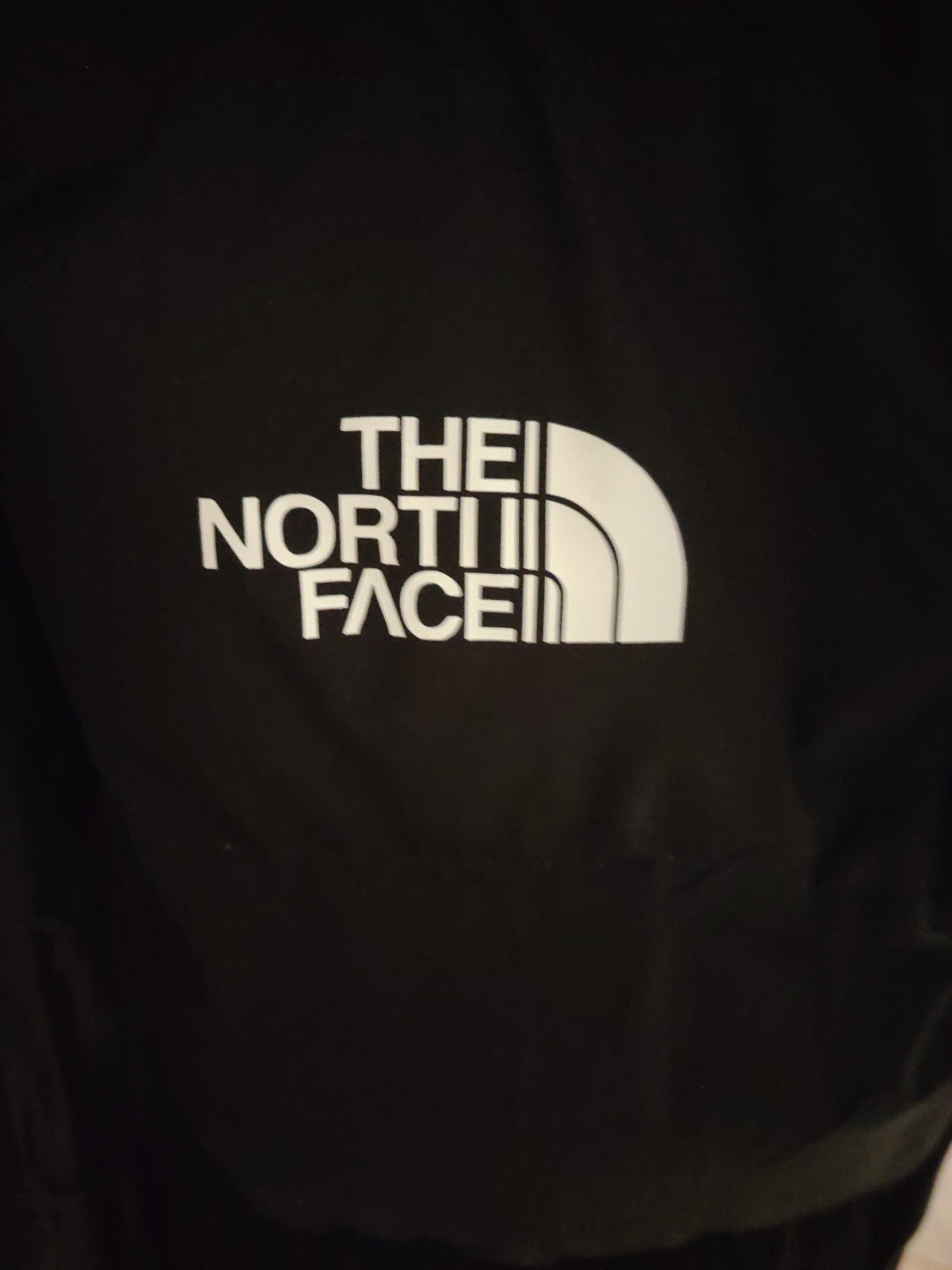 A close-up, angled shot captures the white "THE NORTH FACE" logo prominently displayed on a black fabric garment, likely a jacket or similar apparel, within Huntingdonshire, United Kingdom. The logo features "THE" stacked above "NORTH" which is stacked above "FACE," all in sans-serif capital letters. To the right of the text, the distinctive white half-dome logo is visible. The black fabric background shows subtle texture and minor creasing, indicating a soft, pliable material. The lighting highlights the crispness of the white logo against the dark background. No individuals are present in the frame.
