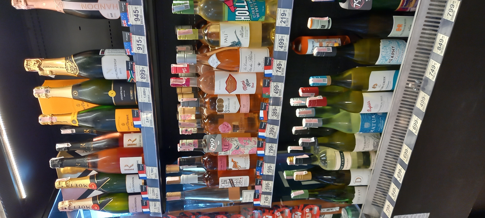 The image captures an indoor retail display of alcoholic beverages, predominantly wine and champagne, in what appears to be a supermarket or liquor store in Moo 1, Thailand. The photo is rotated clockwise, showing shelves filled with bottles arranged vertically.
Numerous bottles of champagne and sparkling wine are visible, including "CHANDON" (Product of Australia) priced at 945.00 and 915.00, "NEETHLINGSHOF BRUT" (Product of South Africa) at 899.00, "SELFRIDGES CHAMPAGNE BRUT" (Product of France) at 1,699.00, "Veuve Clicquot BRUT" (Product of France) at 2,410.00, and "MOET & CHANDON IMPERIAL BRUT" (Product of France) at 2,230.00 and 2,780.00.
The shelves also feature a wide selection of still wines, including white and rosé varieties. Notable brands and prices include "HOLLAND CRISPY WHITE" at 219.00, "YALI wild Swan Sauvignon Blanc" at 499.00, "Les Cerisiers Cotes du Rhône" (Product of France) at 749.00, "Mini Kiss Rosé DA MOSCATO" (Product of Spain) at 499.00, "TRES MEDALLAS" (Product of Chile) at 389.00, "GALETIS" (Product of Chile) at 399.00, "Tops PALE" (Product of USA) at 799.00, and "MATUA" (Product of New Zealand) at 699.00. Other visible brands include "WILD HOUSE," "COBS CREEK," "HEAPHY," and "MAUTHASAR HESS RIESLING."
Each bottle is accompanied by a price tag displaying a three-digit or four-digit number followed by "00," likely indicating Thai Baht, and many feature small country flags denoting the product's origin (e.g., Australia, South Africa, France, Chile, USA, New Zealand, Spain, Holland, Italy, Germany). The scene is well-lit by overhead fluorescent lighting, indicating a commercial setting. No people are visible in the frame. The event taking place is simply the display of products for sale.
