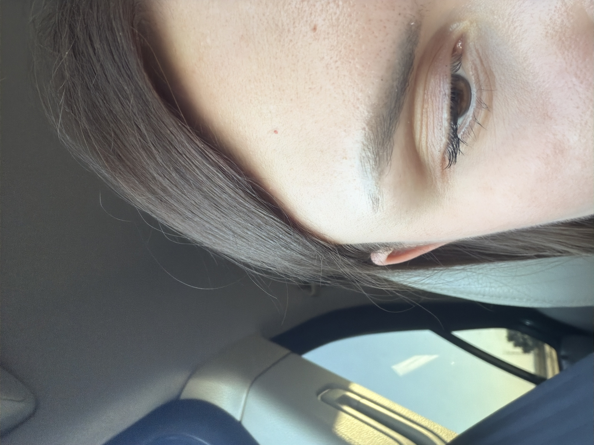 The image shows a close-up of the right side of a person's face, focusing on their eye, eyebrow, forehead, and ear. The person has brown hair and light-colored skin. The eye is open, with visible eyelashes and iris. There are some small red marks on the forehead. The background appears to be the interior of a car, with a window showing a blurry outdoor scene with a clear sky. The image is taken from a slightly upward angle. There is no information about the city of Dolná Streda, Slovakia in the image.