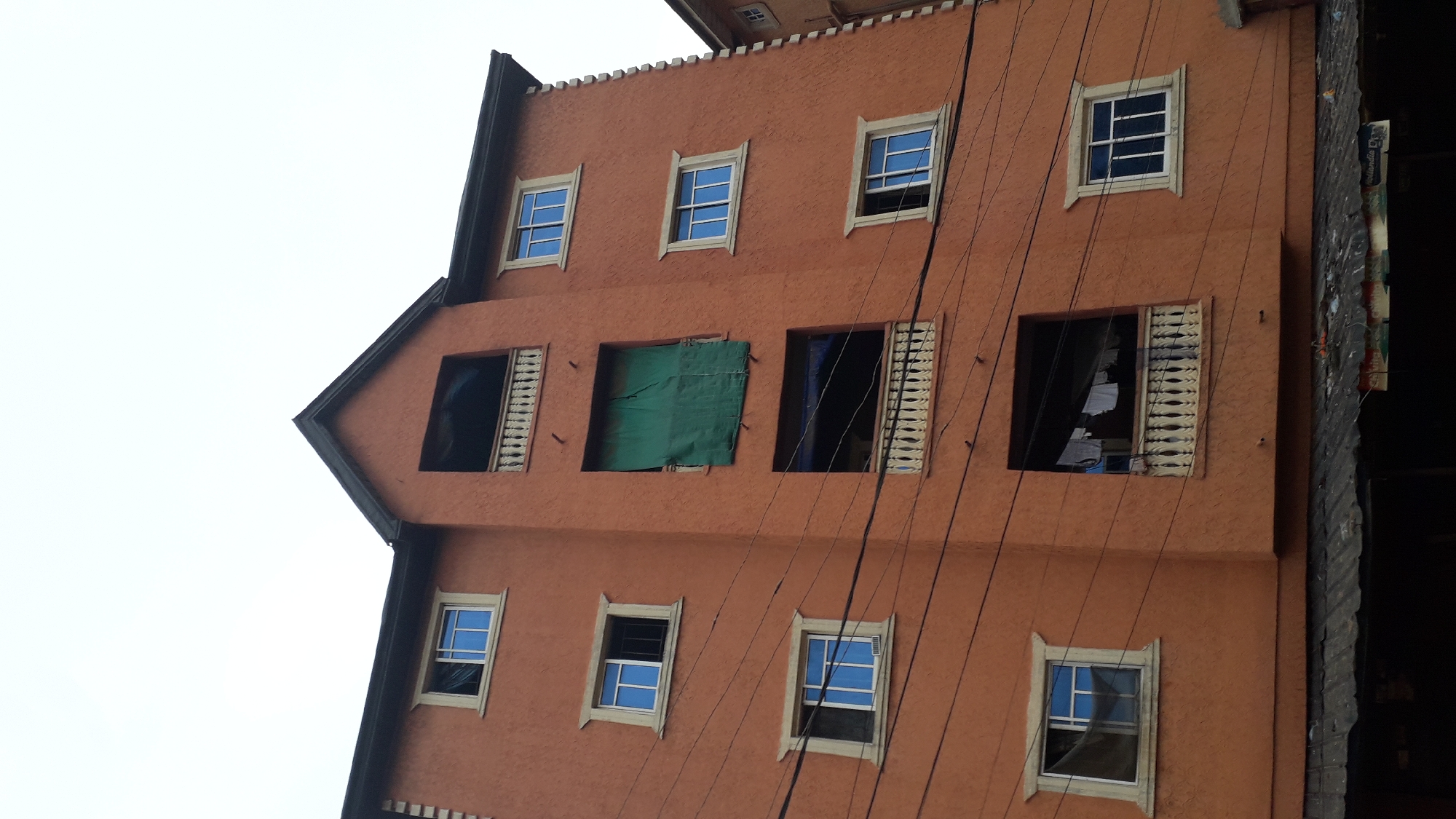 An eye-level, upward-angled shot shows the exterior of a multistory peach-colored building with multiple windows, located in Nkpor, Nigeria. The building features a gabled roofline. The windows on the second level are partly open. The second from the left has a green covering, while the window to the right is draped in a blue-ish cloth. Clothes can be seen hanging in the right-most window on the second floor. The windows are bordered with light trim. Numerous electrical wires crisscross the facade. An adjacent darker building sits partially visible to the right.
