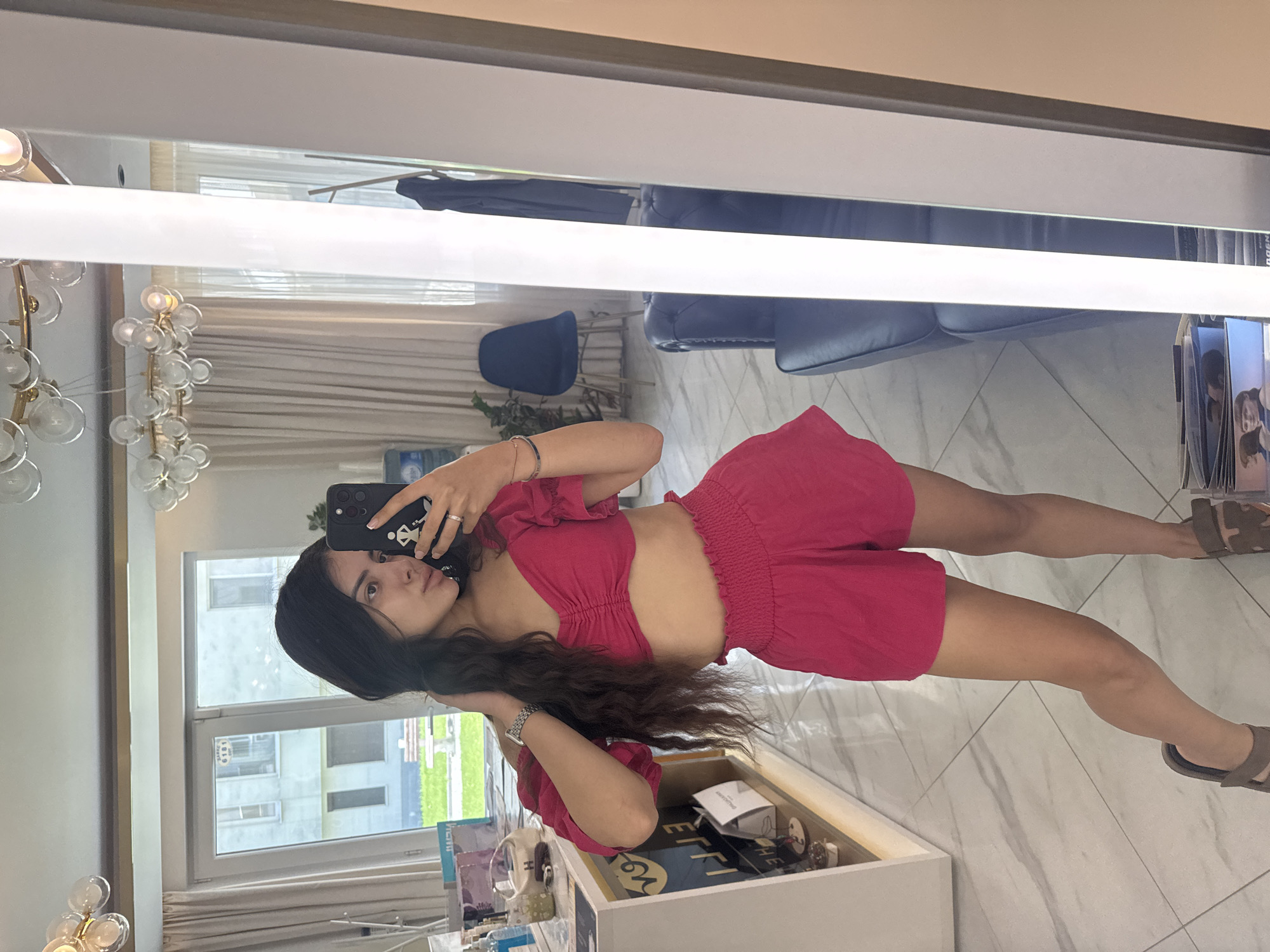 A woman with long, dark hair is taking a mirror selfie in what appears to be an indoor dressing area or showroom. She is wearing a bright pink off-the-shoulder cropped top and matching high-waisted shorts. Her left hand is touching her hair, and her right hand is holding a smartphone, which is angled to capture the reflection. The background shows a large window with a view of a building and some greenery, a modern chandelier, a large mirror with integrated lighting, and a dark blue velvet sofa. To the left, there is a white display unit with various items, including bottles and what looks like merchandise. The floor is tiled with a marble pattern. The scene suggests a contemporary interior space, possibly a boutique or a well-decorated room in a residence. The image does not provide any specific information to place it in Plovdiv, Bulgaria.