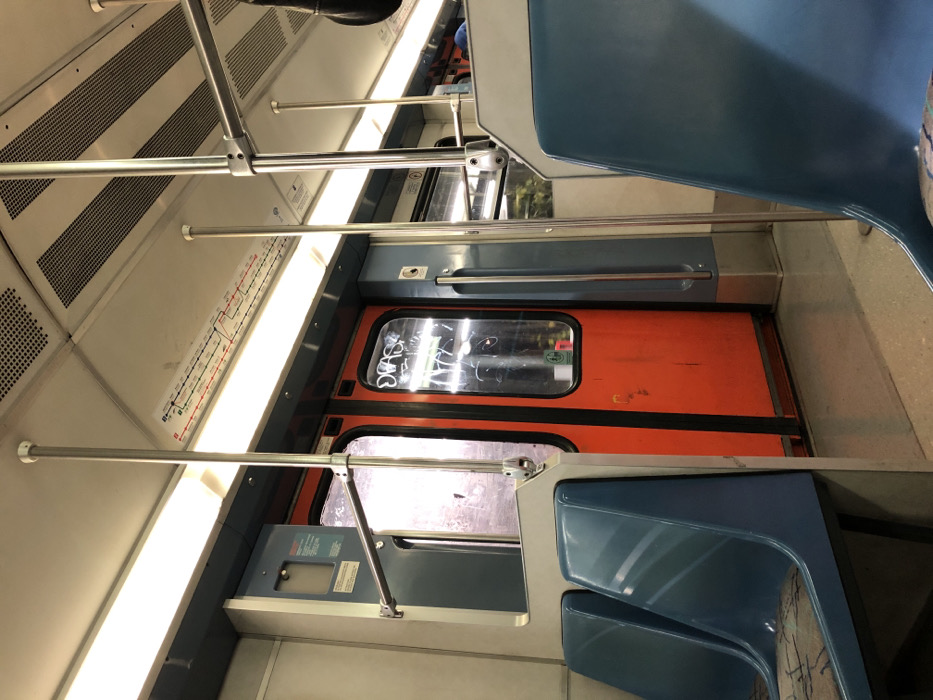 The image displays the interior of a public transport vehicle, likely a metro or train car, in Kallithea, Greece. The photograph is taken from a tilted perspective, showing the length of the carriage.
The scene is devoid of people. Key objects include light blue molded seats, several metal handrails and vertical support poles for passengers, and overhead rectangular light fixtures illuminating the space. The ceiling and upper walls are a light, possibly beige or off-white, color, featuring ventilation grilles.
A prominent orange train door is visible towards the far end of the car. This door has a rectangular window, which is covered with white graffiti or markings, including what appears to be the word "SWAG" and other unidentifiable scribbles. Reflections from the interior are visible on the window glass. A small green sticker is also affixed to the door below the window. Next to the door, a grey panel with some indistinct text or symbols is present.
Along the ceiling, a linear diagram, likely a metro or train route map, is displayed, showing a series of interconnected stations. However, the text on this map is too small and blurry to be deciphered. The floor is light-colored and appears to have a textured or patterned surface.
No specific activity or event is taking place, as the car is empty. The interior lighting suggests it could be daytime or night, but no external view is available to confirm. No emotional cues are present.