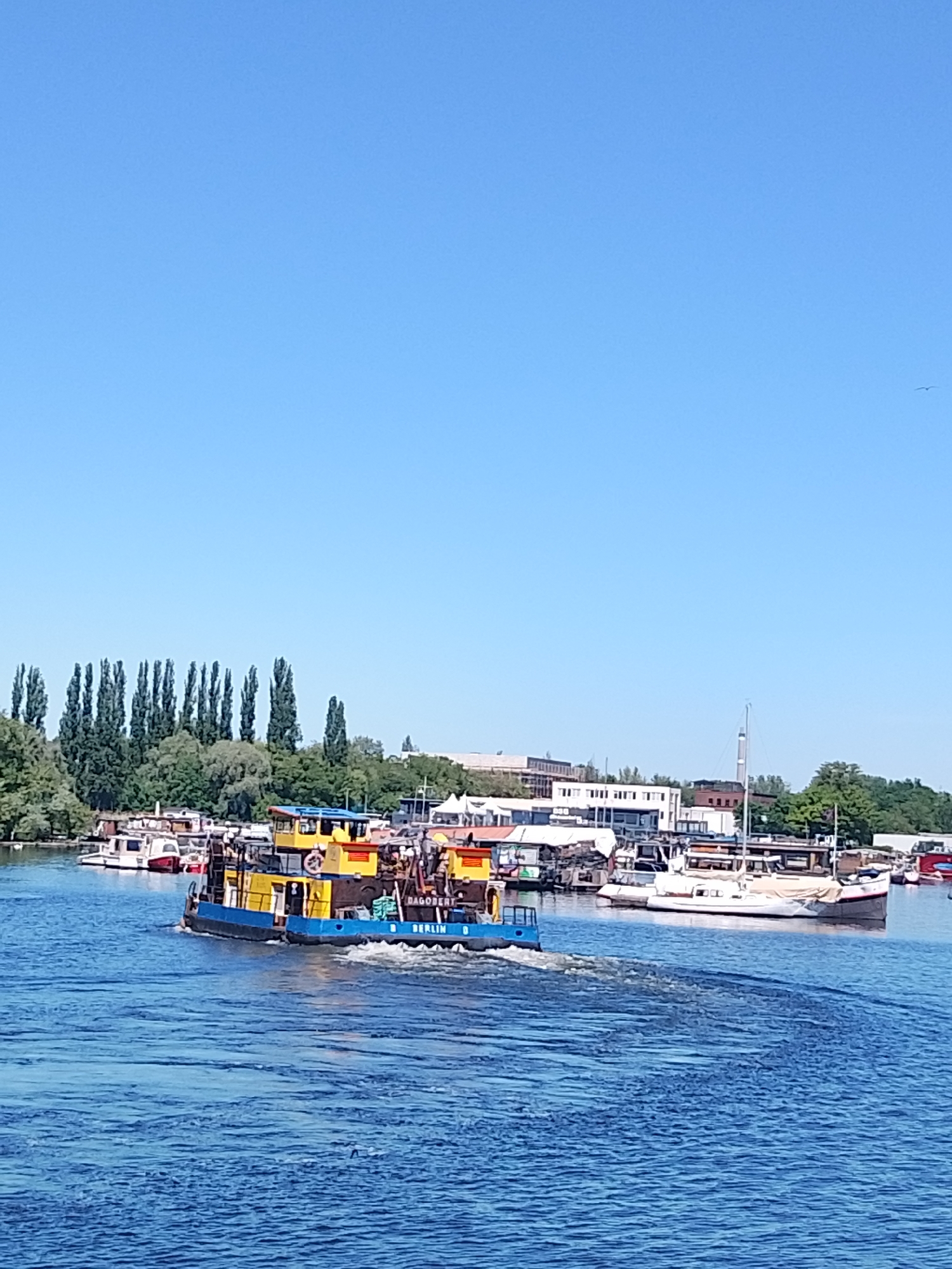 The image depicts a waterfront scene. A blue and yellow tugboat with the name "BERLIN D" is moving through the water, leaving a wake behind it. In the background, there are various boats docked along the shore. A building complex is visible behind the boats, with trees interspersed. The sky is clear blue. The location is in the city of Unknown, Unknown.