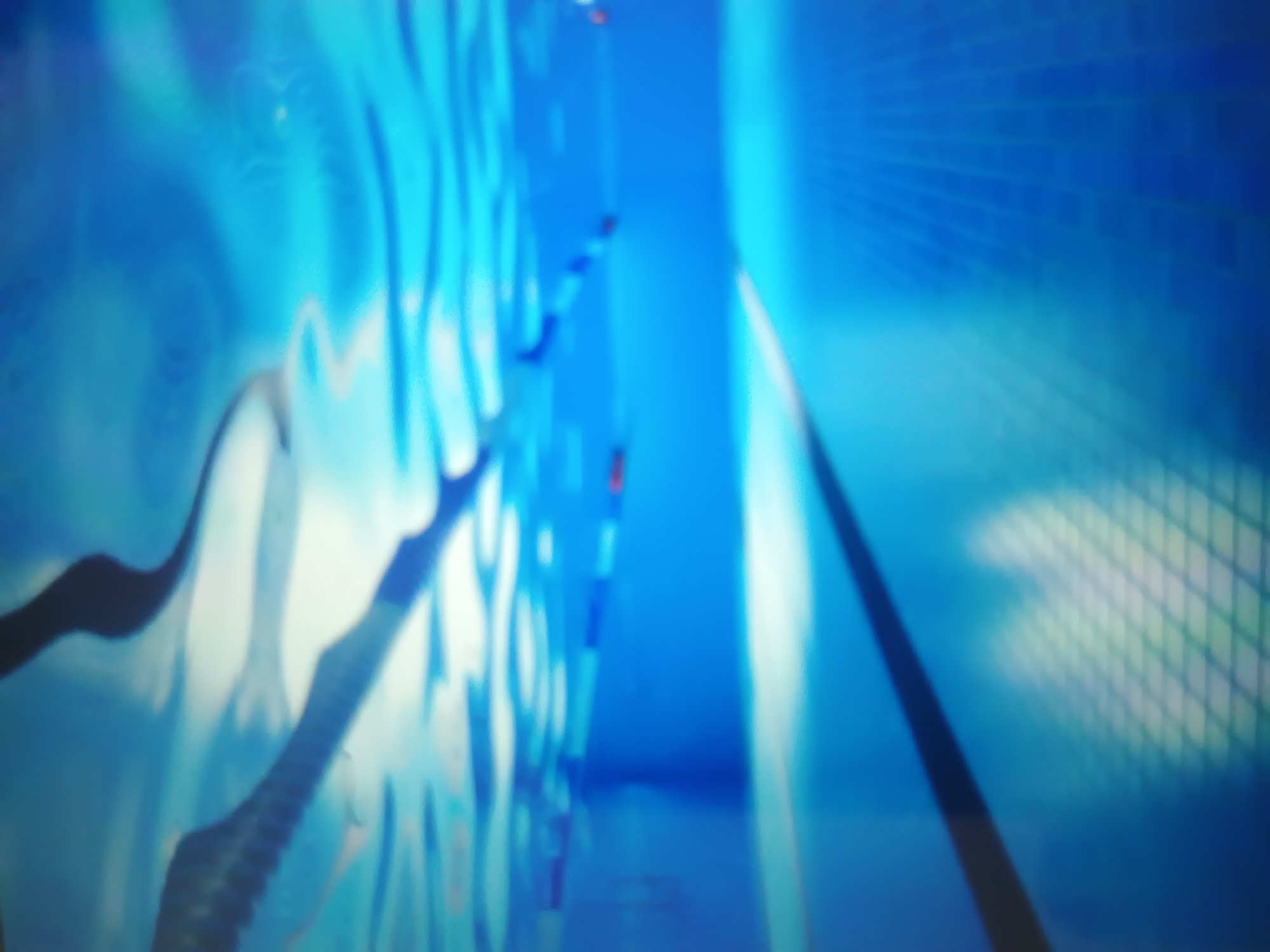 This image presents an underwater perspective within a swimming pool, situated in Kroměříž, Czechia. The clear blue water occupies the foreground and midground, with a visible, rippled water surface above, exhibiting strong light reflections and refractions. Dark, blurred shapes, consistent with lane ropes and their shadows, are cast against the bright surface. Multiple lane ropes, featuring segments of red and blue, traverse the pool horizontally, both above and submerged in the water. The pool walls are uniformly blue and extend into the background. The tiled pool floor, adorned with a discernible grid pattern, stretches from the foreground towards the distant end, centrally marked by a dark lane line. No individuals are present in the scene.