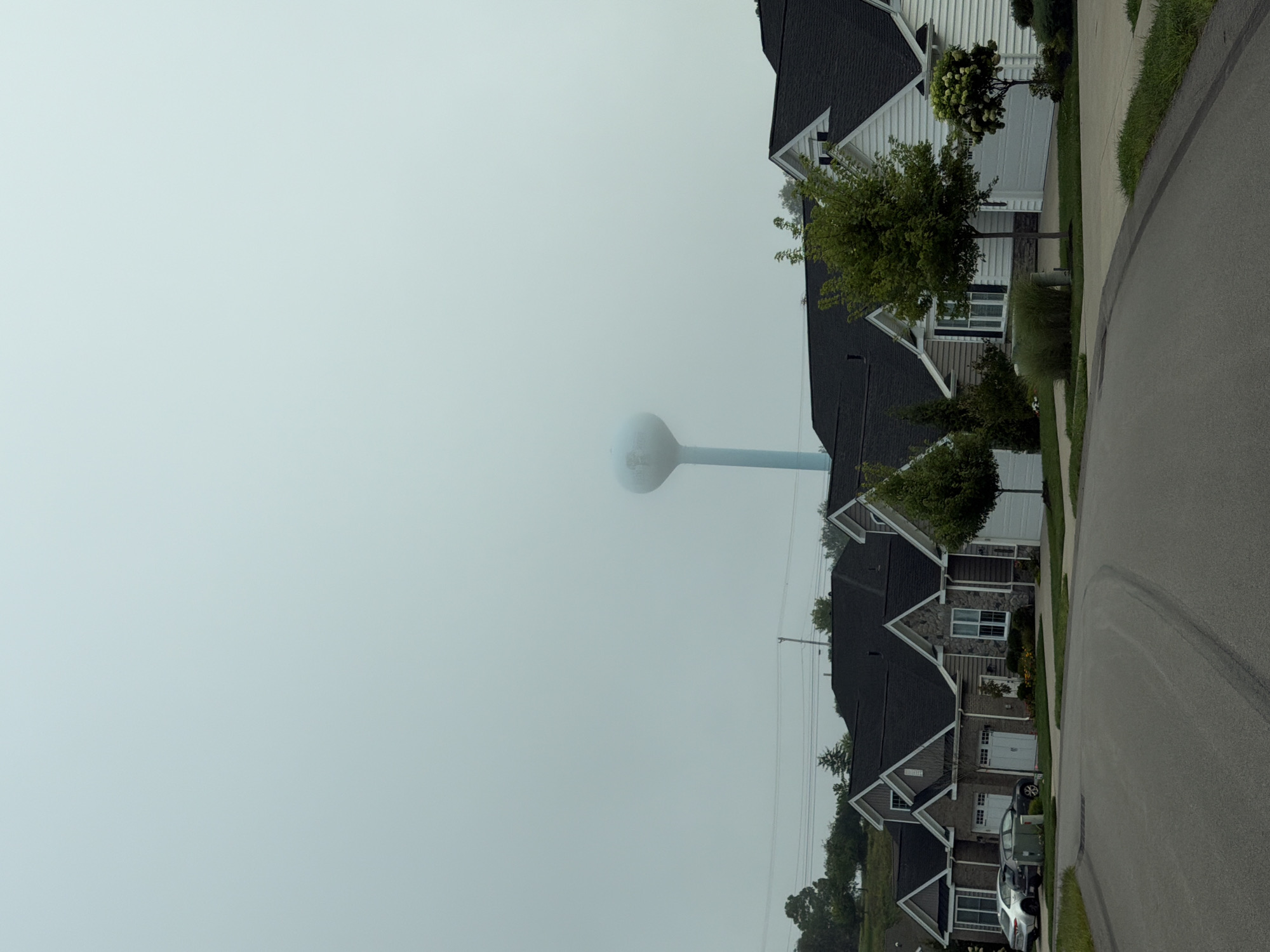 The image depicts a suburban street scene in Richland Township, United States of America, under a thick, hazy sky. A row of modern houses with dark roofs and light-colored siding lines the right side of the frame. Some houses feature stone accents and multiple garage doors. Landscaping, including trees and bushes, is visible in front of the houses. In the foreground, an asphalt road curves to the left, with a paved driveway branching off to the right. Two cars are parked in front of the houses on the left. In the background, a tall, light-blue water tower stands prominently, its top partially obscured by the haze. The overall atmosphere is overcast and somewhat muted due to the weather conditions.