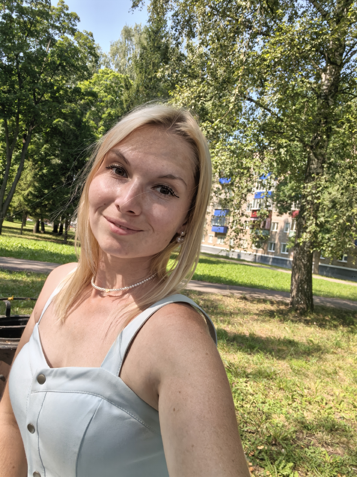 A medium shot, outdoor, selfie-style photograph captures a young Caucasian woman with blonde hair and light-colored eyes smiling slightly at the camera. She is wearing a light blue sleeveless dress with visible buttons down the front and a pearl necklace with a small heart-shaped pendant. She is positioned in the foreground, with her upper body and face prominently featured.
The background suggests a park-like setting on a sunny day. Lush green trees with dappled sunlight are visible behind her, with a paved path and a grassy area further back. A multi-story apartment building with colorful balconies is partially visible through the trees on the right side of the frame. The location is identified as Sterlitamak, Russia, indicating an urban or suburban park environment. The overall atmosphere is casual and relaxed.