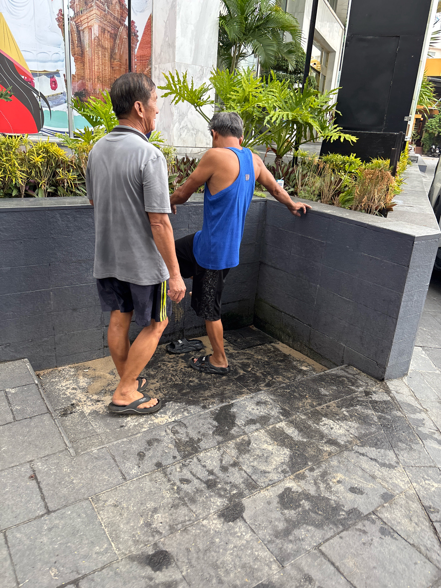 Two middle-aged Asian men are standing in an outdoor urban setting, possibly Nha Trang, Vietnam, near a planter box with lush green foliage. The man on the left is wearing a grey t-shirt and dark shorts with yellow stripes. He is facing away from the camera, looking towards the man on the right. The man on the right is wearing a blue tank top and black shorts. He is leaning against a dark grey, tiled wall, with his right leg extended, pressing against the wall. His left arm is resting on the top of the wall. Both men are wearing flip-flops. The ground is paved with grey tiles and appears wet and dirty in patches, suggesting recent rain or cleaning. In the background, a large mural depicting a temple-like structure is visible. A large black rectangular object, possibly a sign or part of a building, is also present. The overall impression is that of a casual encounter in a public space.