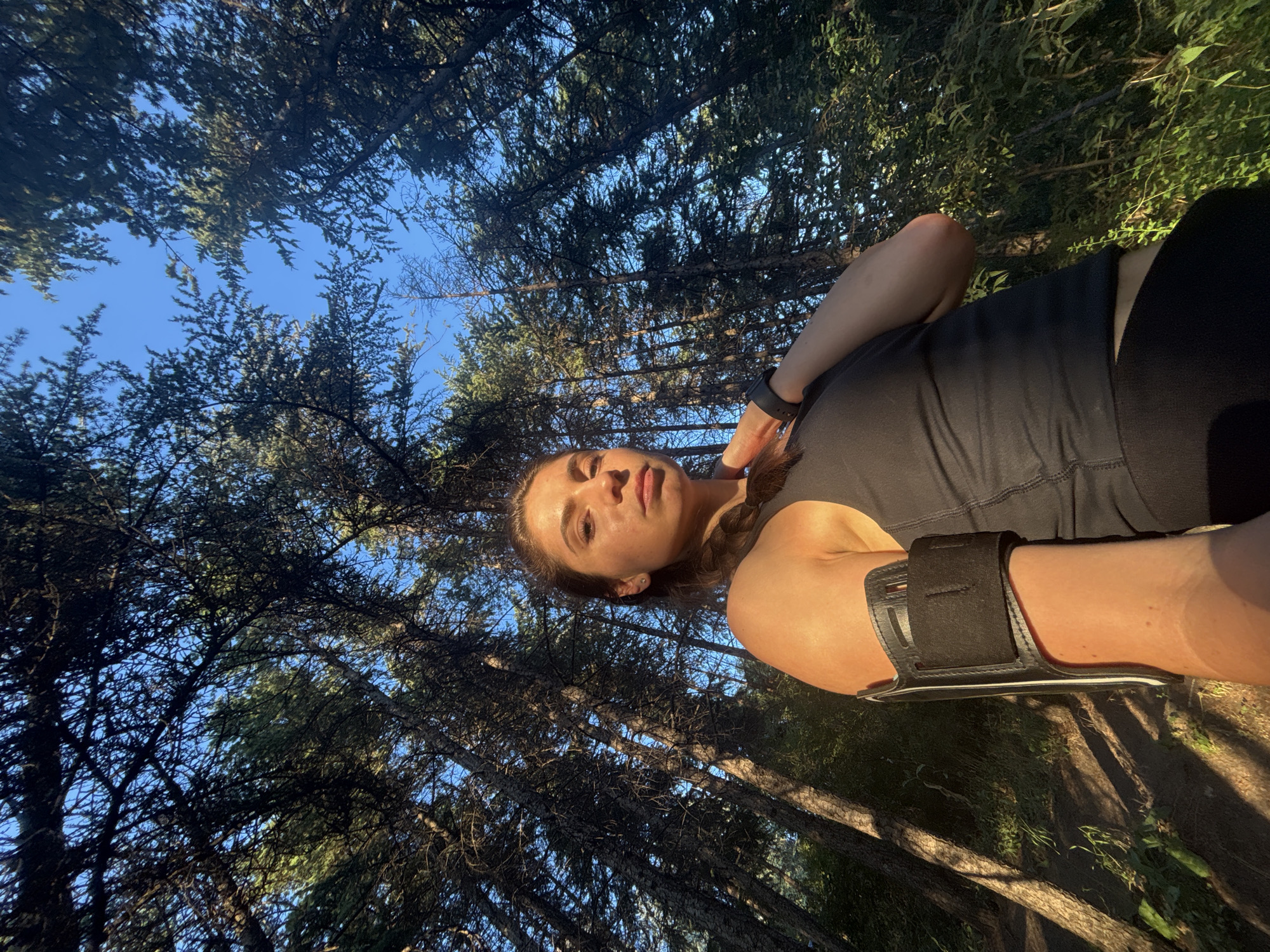 The image captures a low-angle, wide shot of a young woman in a forest setting, looking up at the camera. She is wearing a black tank top and black leggings, and an armband is visible on her left arm. Her gaze is directed towards the camera, and her expression is neutral. Sunlight filters through the dense canopy of pine trees, creating dappled shadows on the forest floor and on her skin. The time of day appears to be late afternoon or early evening, given the warm, golden light and long shadows. The blue sky is visible between the trees, suggesting clear weather. The location is a forest, likely in Varna, Bulgaria, with tall pine trees characteristic of the region. The overall mood is one of outdoor activity, possibly a break during a run or hike, with a sense of being immersed in nature. There is no visible text in the image.