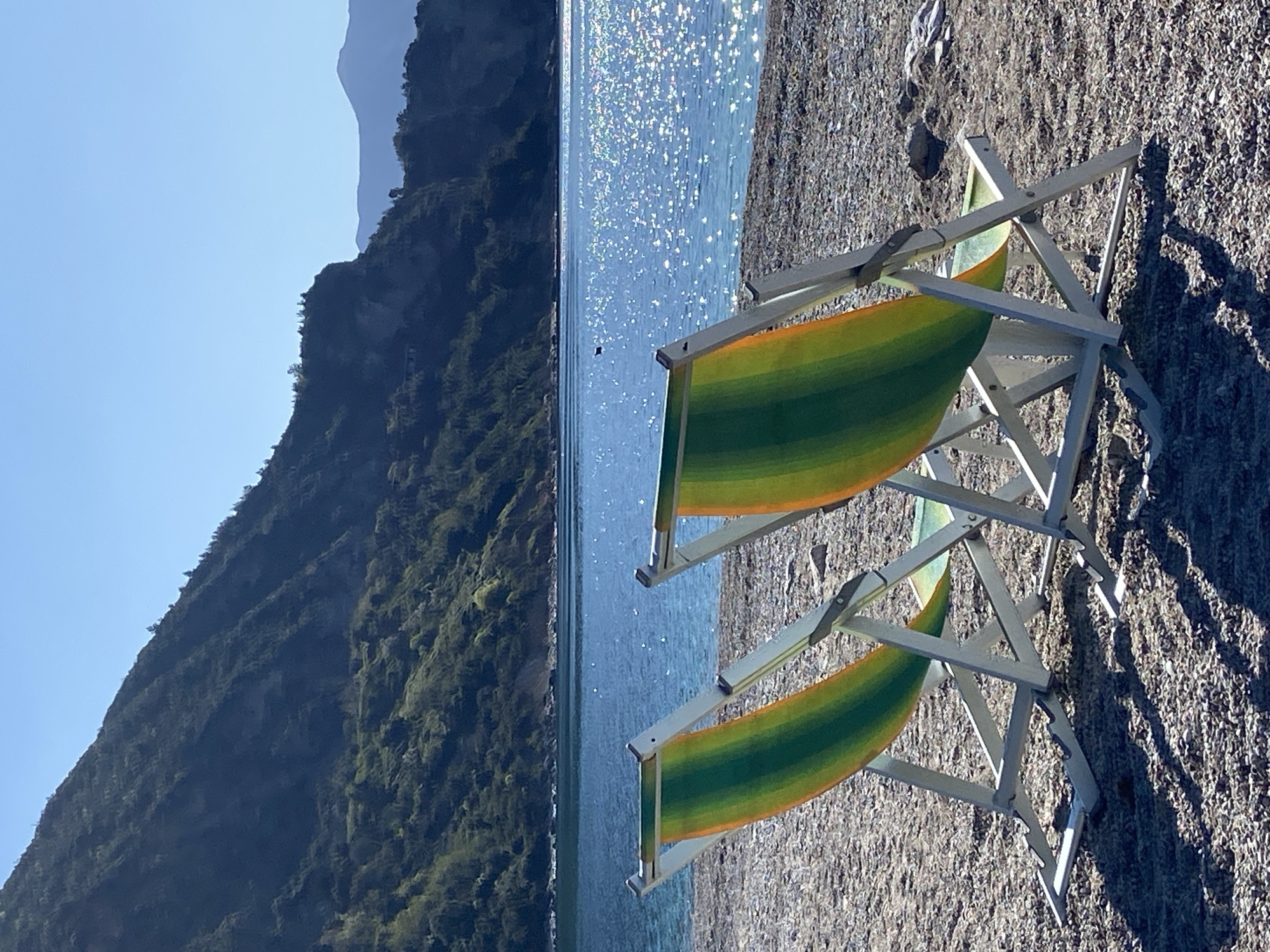 Two beach chairs are positioned on a pebble beach. The chairs have green and yellow striped fabric and white frames. The Adriatic Sea shimmers in the background, reflecting sunlight. A mountain rises on the left side of the frame. The image location is likely near or in Prijevor, Montenegro. There are no people visible in the frame.