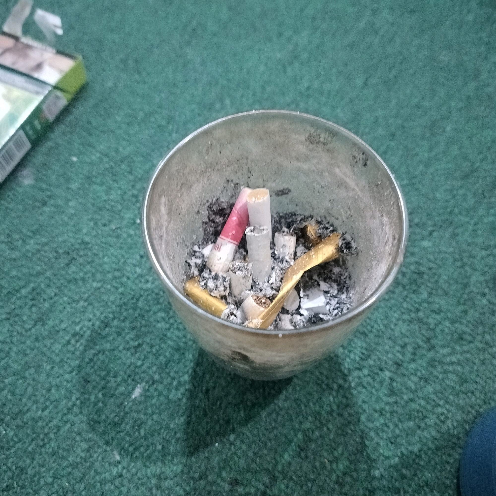 An ashtray filled with cigarette butts and ash sits on a green carpet. A cigarette pack is visible in the upper left corner of the image. The ashtray is made of metal and appears to be old and dirty. The cigarette butts are of various types, some with filters and some without. There is also some loose ash and debris in the ashtray. The image is taken from a high angle, looking down into the ashtray. The lighting is dim and the image is slightly blurry. The scene is consistent with a typical indoor setting in Ngemplak, Indonesia.