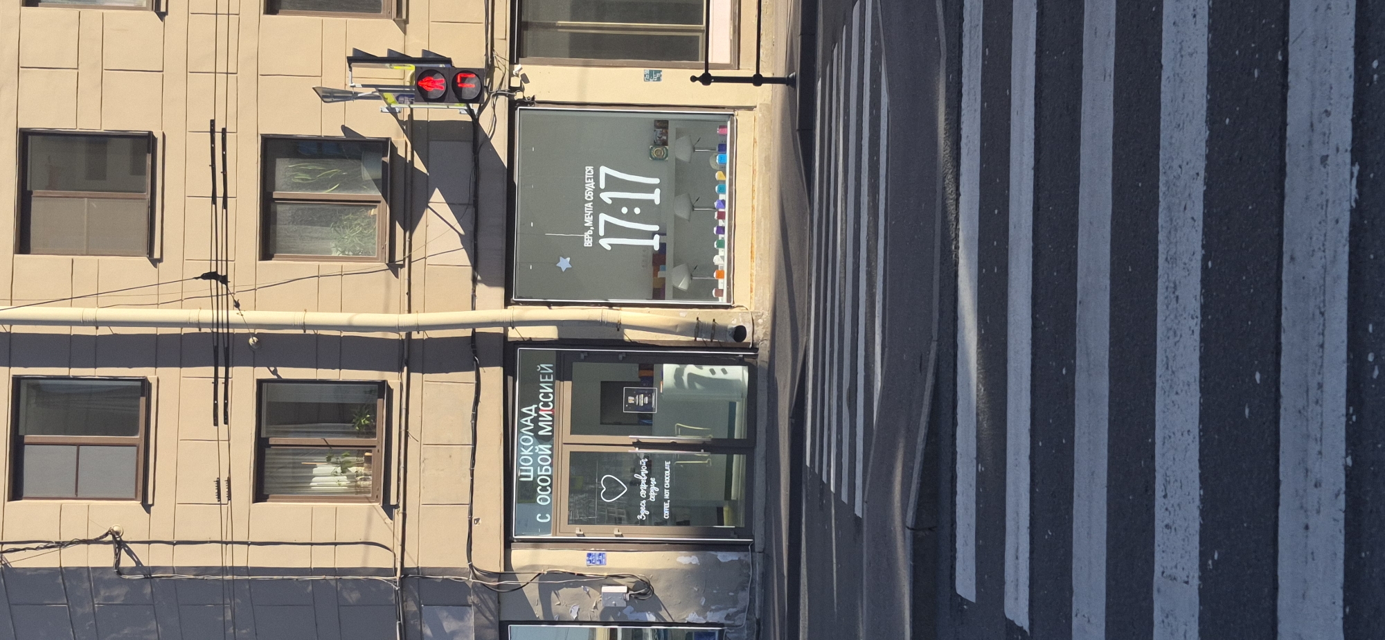 The image shows a street scene in Saint Petersburg, Russia. In the foreground, a pedestrian crossing with white stripes is visible. Adjacent to the crossing is a building with several windows. One window displays a digital clock showing "17:17" and the Russian text "ВЕРЬ, МЕЧТА СБУДЕТСЯ," which translates to "Believe, the dream will come true." Below this, another shop window has text in Russian, "ШОКОЛАД С ОСОБОЙ МИССИЕЙ," meaning "Chocolate with a Special Mission." Inside this lower window, a heart symbol and text indicating "COFFEE, HOT CHOCOLATE" are visible. A traffic light with a red signal is also mounted on the building. The lighting suggests it is daytime, likely late afternoon, given the time displayed on the clock and the long shadows cast by the building and traffic light. The overall scene depicts an urban environment with commercial establishments and public infrastructure.