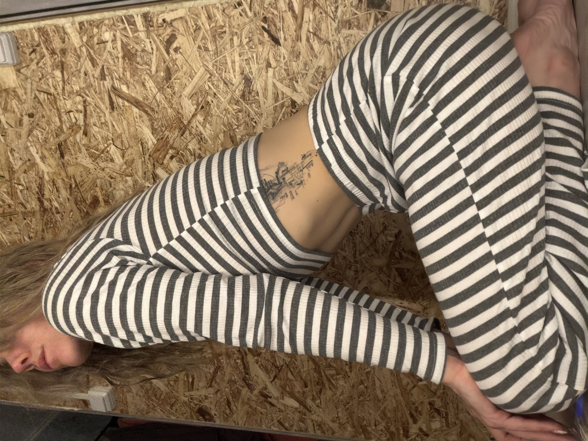 A female individual is depicted kneeling in an indoor setting in Chaikovsky, Russia. She is positioned in a left profile view, with her head tilted back, eyes obscured or closed, and long, light-colored hair cascading over her shoulders. She wears a two-piece ribbed knit outfit consisting of a long-sleeved crop top and matching pants, both featuring horizontal stripes of alternating dark grey and white. A black tattoo, appearing as an intricate, schematic-like design, is visible on her left lower back/flank. Her hands are placed together in the lower-left foreground on a surface not fully visible, and her feet are bare. The background consists of a wall covered with Oriented Strand Board (OSB) panels, characterized by visible wood chips. A white electrical fixture, possibly a switch or outlet, is partially discernible on the OSB wall in the upper-left quadrant.