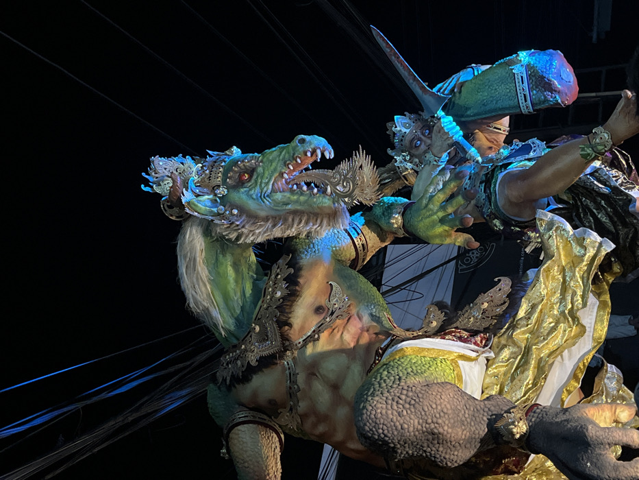 The image displays two large, elaborate effigies illuminated at night in Canggu, Indonesia. The dominant subject is a colossal, bipedal figure, likely an Ogoh-Ogoh, with a reptilian-like head, green scaly skin, and a muscular, human-like torso. Its face features glowing red eyes, prominent teeth, and a white and green mane, adorned with an ornate metallic silver-gold crown. Similar silver-gold decorations embellish its chest, shoulders, and arms. Its lower body is draped in rich gold and white fabric.
Below and to the right of this large figure, a smaller, human-like effigy is depicted in a crouched or struggling posture. This smaller figure also wears an ornate crown and armbands, possesses bright blue eyes, and exhibits greenish skin tones. It appears to be holding a long, pointed, blade-like object. Both figures are illuminated by artificial lights, casting a prominent blue glow, particularly on the faces and lower extremities, with some warm yellow light highlighting the larger figure's chest and gold fabric.
The background is a dark, starless night sky. Multiple thin, dark wires or cables crisscross the upper portion of the frame. A dark banner with white text, partially legible as "KOKOS VILLA" and a web address, is visible in the mid-ground behind the effigies. The scene captures a dynamic interaction between the towering, reptilian figure and the smaller, human-like figure.