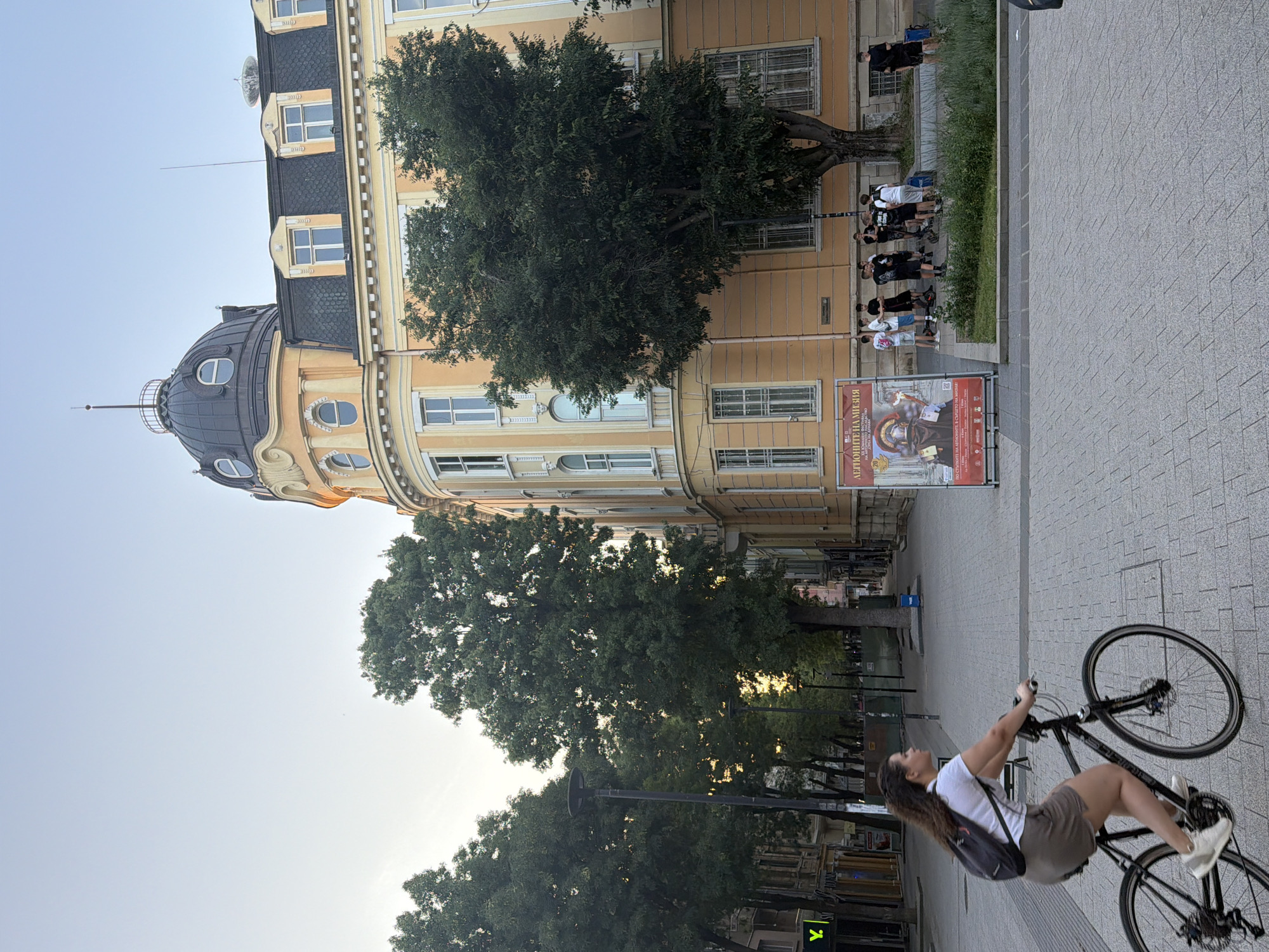 The image captures a street scene in Pleven, Bulgaria, likely in the city center given the architecture and pedestrian activity. In the foreground, a young woman is riding a bicycle to the left, wearing a white t-shirt, brown shorts, and a backpack. She is on a paved sidewalk. To her right, a large, ornate building with a yellow facade and a dark, domed roof dominates the background. The building has multiple windows, some arched, and architectural detailing. In front of the building, a group of young people are gathered, some standing and some with electric scooters. A large poster advertising an exhibition is also visible near the building. Trees line the street, casting shadows, and streetlights are present. The sky is a pale blue, indicating it's likely late afternoon or early evening.
