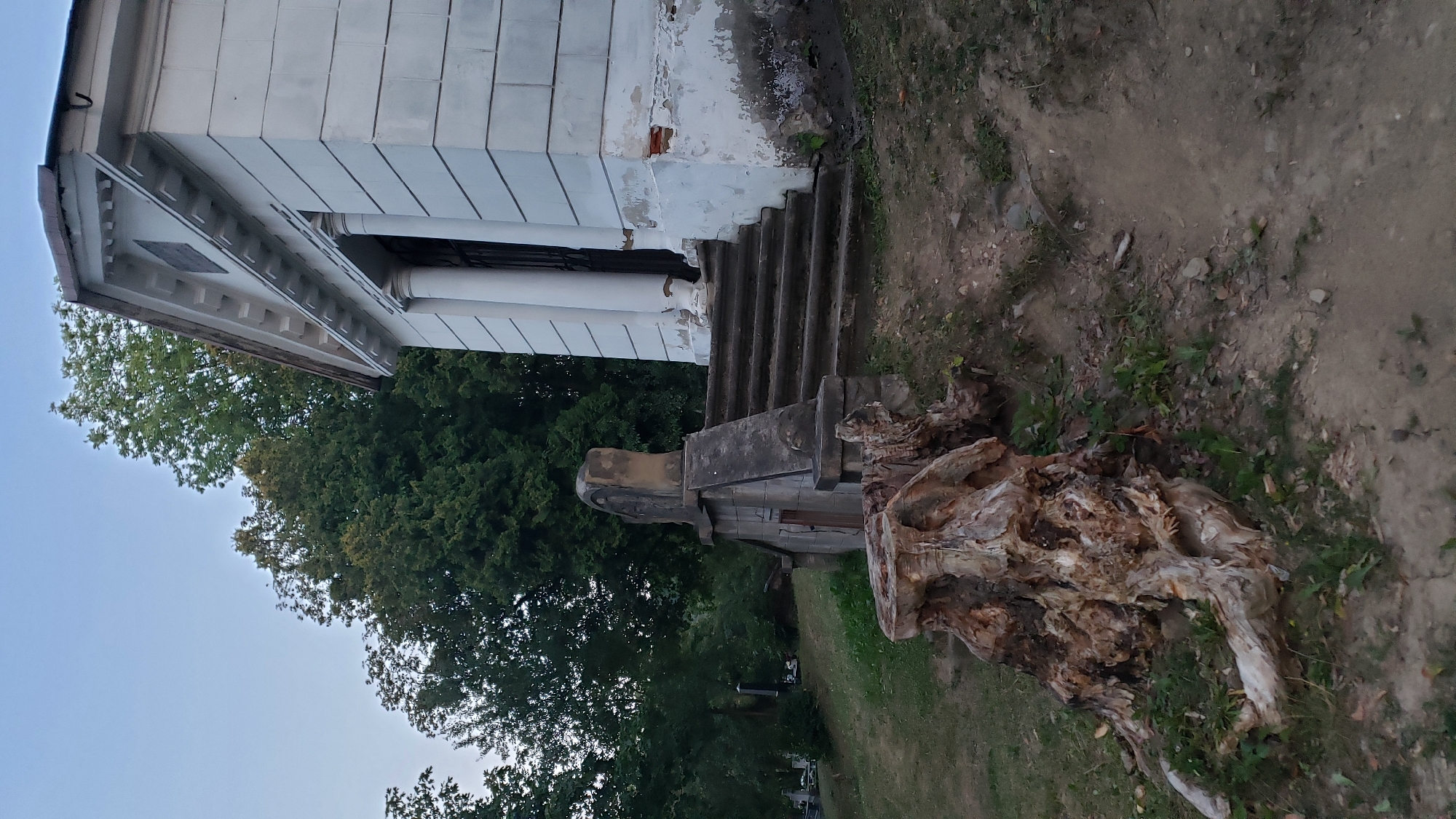 The image depicts a scene in the cemetery of Prešov, Slovakia. In the foreground, a large, gnarled tree stump is visible on the left, with exposed roots and weathered wood. Behind the stump, a stone structure with a small door and a weathered stone monument on top is situated on a grassy slope. To the right of this structure, a set of stone stairs leads up to a white building with classical columns and a pediment. The building appears to be a mausoleum or chapel, with signs of peeling paint on its lower facade. Lush green trees form a backdrop, and the sky is a pale blue, suggesting it is either early morning or late evening. The ground is a mixture of grass, dirt, and fallen leaves.