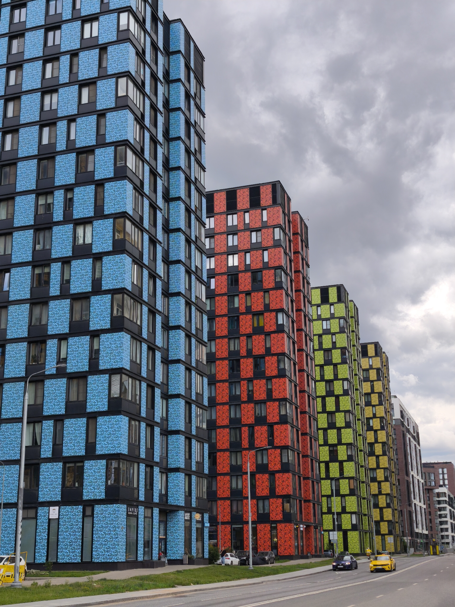 This media file depicts a row of brightly colored, modern apartment buildings in Moscow, Russia. The buildings are multi-story and feature geometric patterns on their facades, with the closest one being predominantly blue and black. Further down the row, the buildings transition to red and black, then lime green and black, and finally yellow and black, creating a vibrant visual spectacle. The scene appears to be a street-level view of the buildings, with a road in the foreground and cars driving past. The sky is overcast and cloudy, suggesting a cooler, perhaps overcast day.
There are a few vehicles visible on the road, including a yellow car in the foreground, a dark blue sedan, and other cars parked or driving in the distance. The presence of streetlights and some sparse greenery along the roadside indicates an urban environment. A few small signs are visible on the lower levels of the blue building, but the text is not clearly legible. The overall impression is one of contemporary urban architecture with a playful and bold design aesthetic, set against a typical cloudy Moscow sky. No specific activity or event is clearly depicted beyond the ordinary flow of urban life, such as traffic. The weather conditions and the cloud cover contribute to a somewhat muted, but still striking, atmosphere.