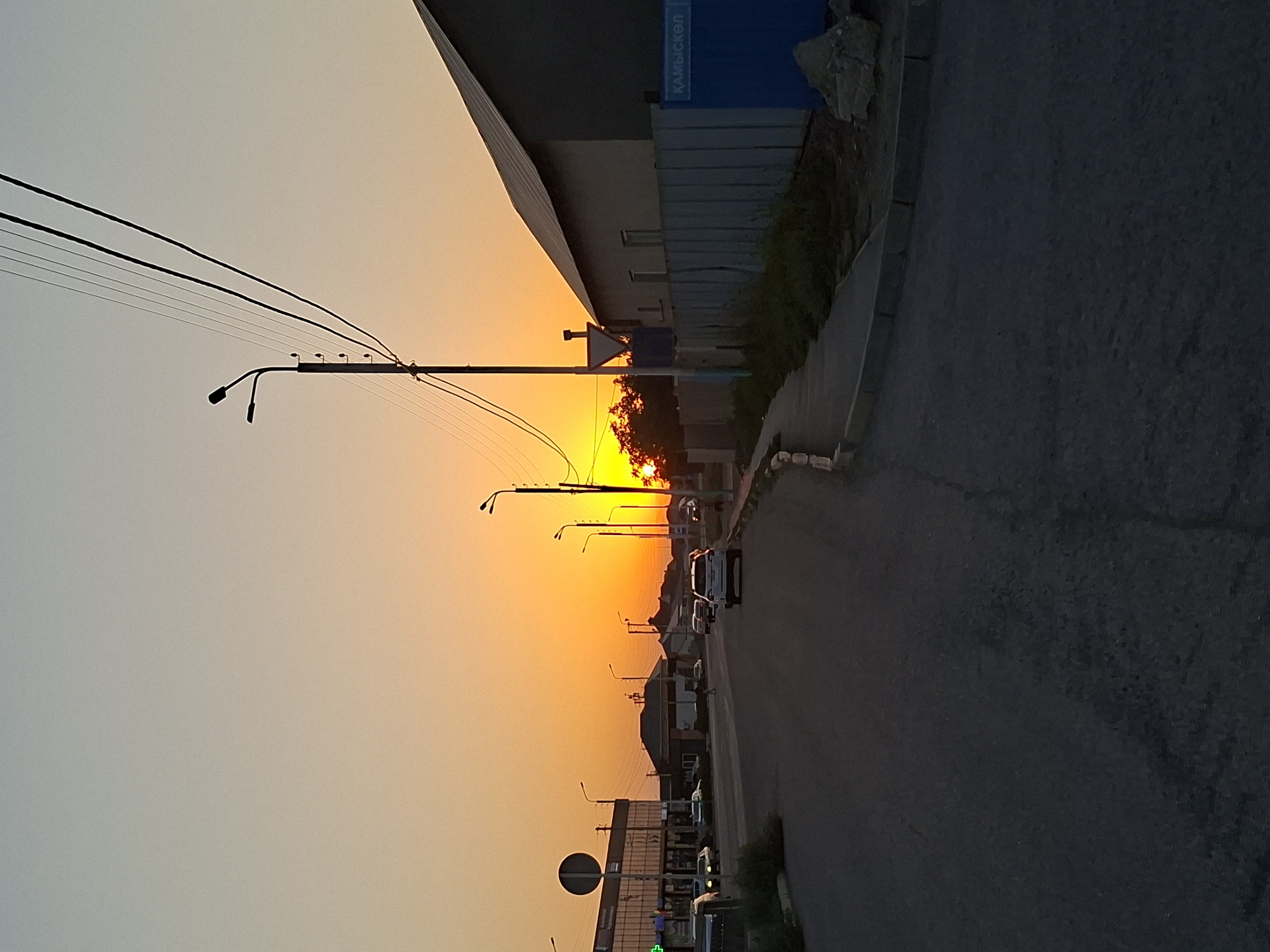 This image depicts a street scene at sunset in Atyrau, Kazakhstan. The sky is dominated by a vibrant orange and yellow glow, indicating the sun is low on the horizon. Silhouetted against this dramatic sky are power lines and streetlights, stretching across the frame.
On the right side of the image, a paved road runs alongside a sidewalk. Several vehicles, including a white van and other cars, are parked or driving along the road. Buildings line the street, some with visible windows and architectural details. One building has a blue sign with white Cyrillic text that appears to read "КАМЫСКЕЛ". A grey fence runs along the edge of some of the properties. Trees with dense foliage are also visible, their leaves catching the warm light of the sunset.
The overall mood of the image is peaceful, with the warm hues of the sunset creating a serene atmosphere. The scene captures a typical evening in an urban or semi-urban environment, with everyday objects like streetlights and vehicles integrated into a beautiful natural phenomenon. The time of day is clearly sunset, and the weather appears clear and calm.
