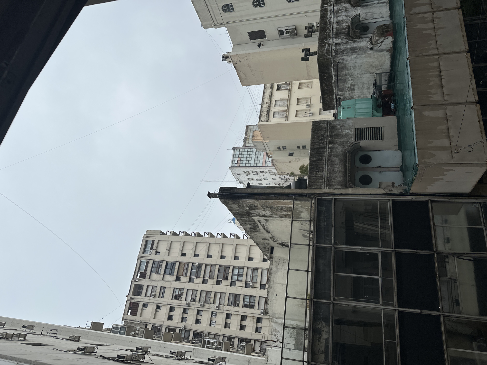 The image depicts a view looking up at buildings in Buenos Aires, Argentina, under an overcast sky. The buildings are varied in style, ranging from white facades with multiple windows and air conditioning units to older, dirtier buildings with large, industrial equipment on their roofs. A cable runs between two of the buildings. In the foreground, there are visible rooftops with vents and air conditioning units.