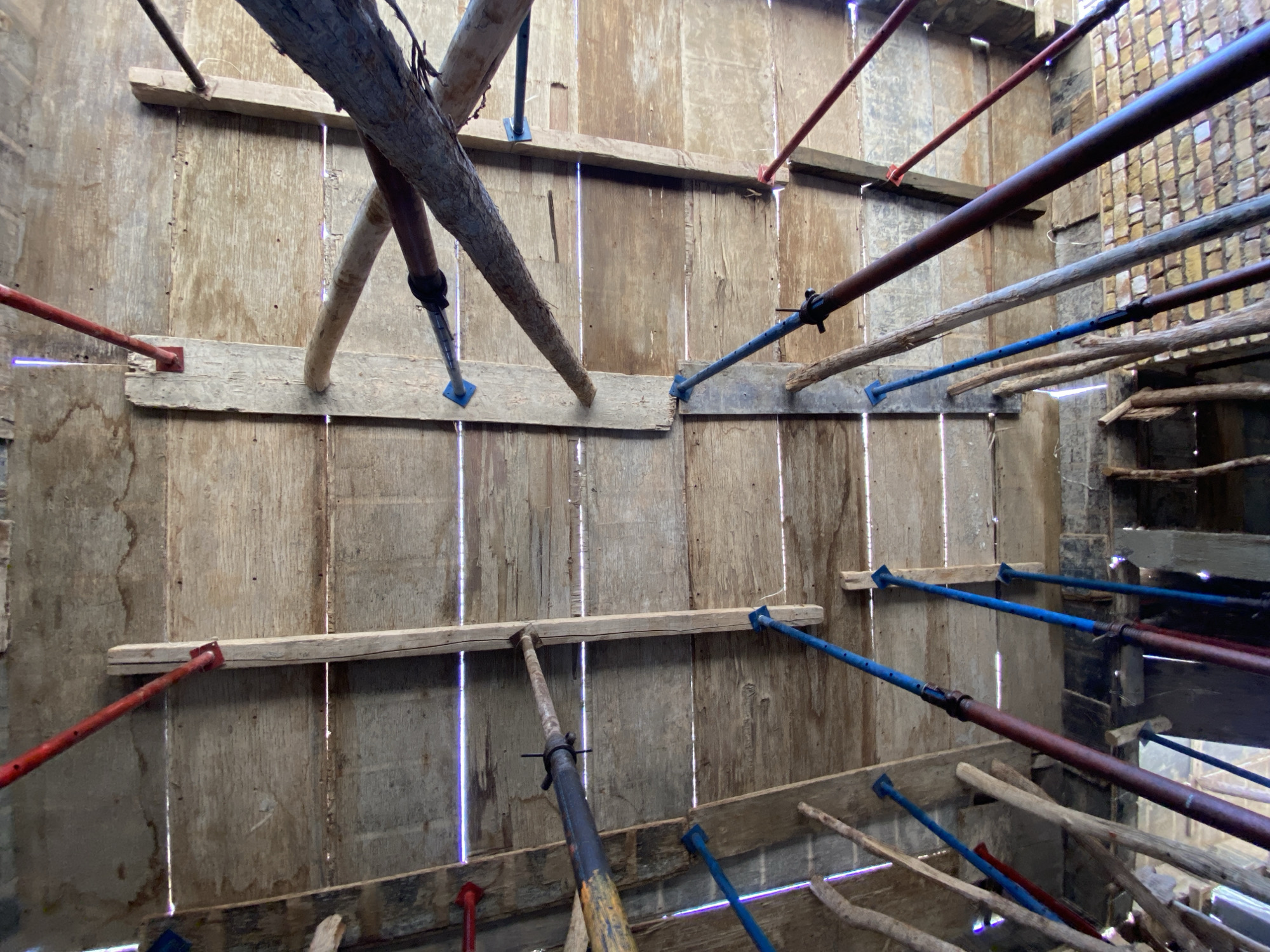 The image depicts a close-up view of the interior of a construction site. The scene includes wooden formwork supported by adjustable metal props, some painted red or blue. The props are wedged against the wooden planks to support the formwork. The wood appears to be a combination of planks and small tree trunks. Vertical gaps are visible between the boards. In the background, a brick wall under construction is partially visible. The image is taken from an indoor location. There is no visual data to associate with Kandahar or Afghanistan, so they are not included in the description.