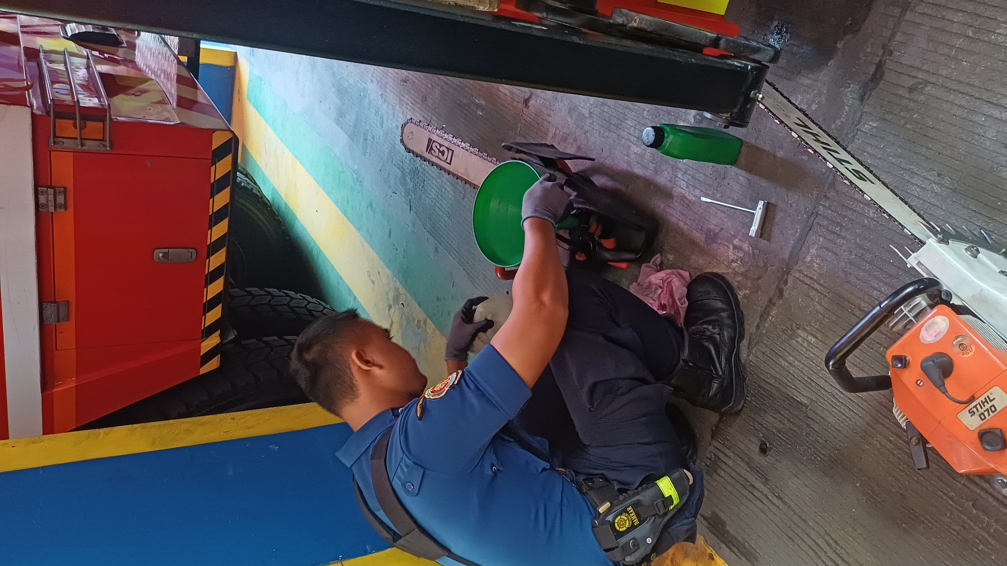 In South Jakarta, Indonesia, a firefighter in a blue uniform and gloves is kneeling on a grey concrete floor, attending to a chainsaw. The firefighter is holding a green funnel over the chainsaw's fuel tank. Next to the chainsaw are two bottles of oil, one green and one white. In the foreground, another orange chainsaw is visible, partially obscured by the first. In the background, a large red vehicle, possibly a fire truck, is parked. The environment appears to be a garage or maintenance area, with yellow and blue markings on the wall and floor.