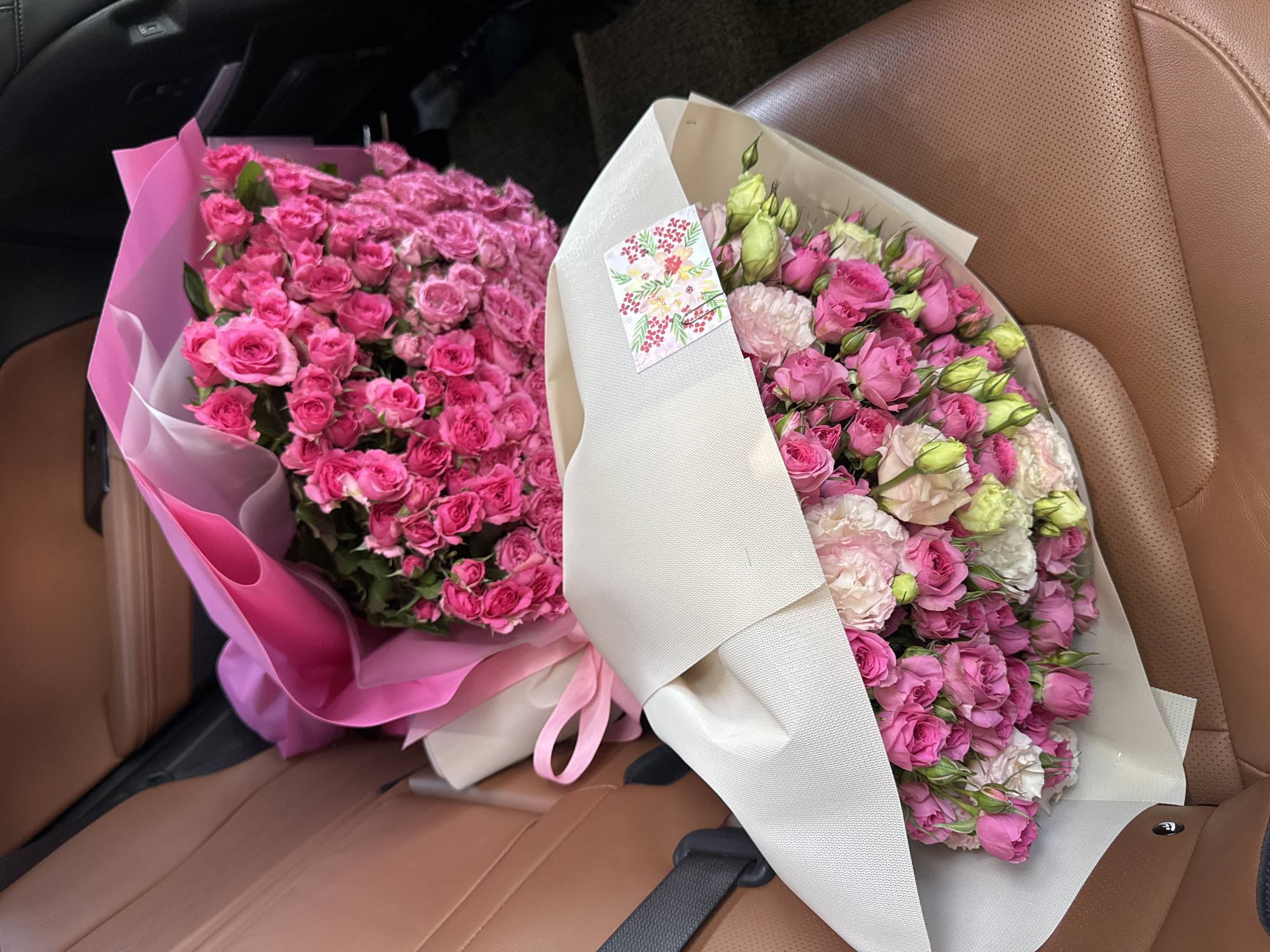 Two bouquets of pink and white roses are arranged on the perforated brown leather seats of a car, likely in Almaty, Kazakhstan. The bouquet in the foreground is wrapped in light-colored paper and features a small card with a floral design. The bouquet behind it is larger and wrapped in vibrant pink paper. A grey seatbelt is visible on the left side of the frame. The car's interior is dimly lit, with the focus on the flowers.