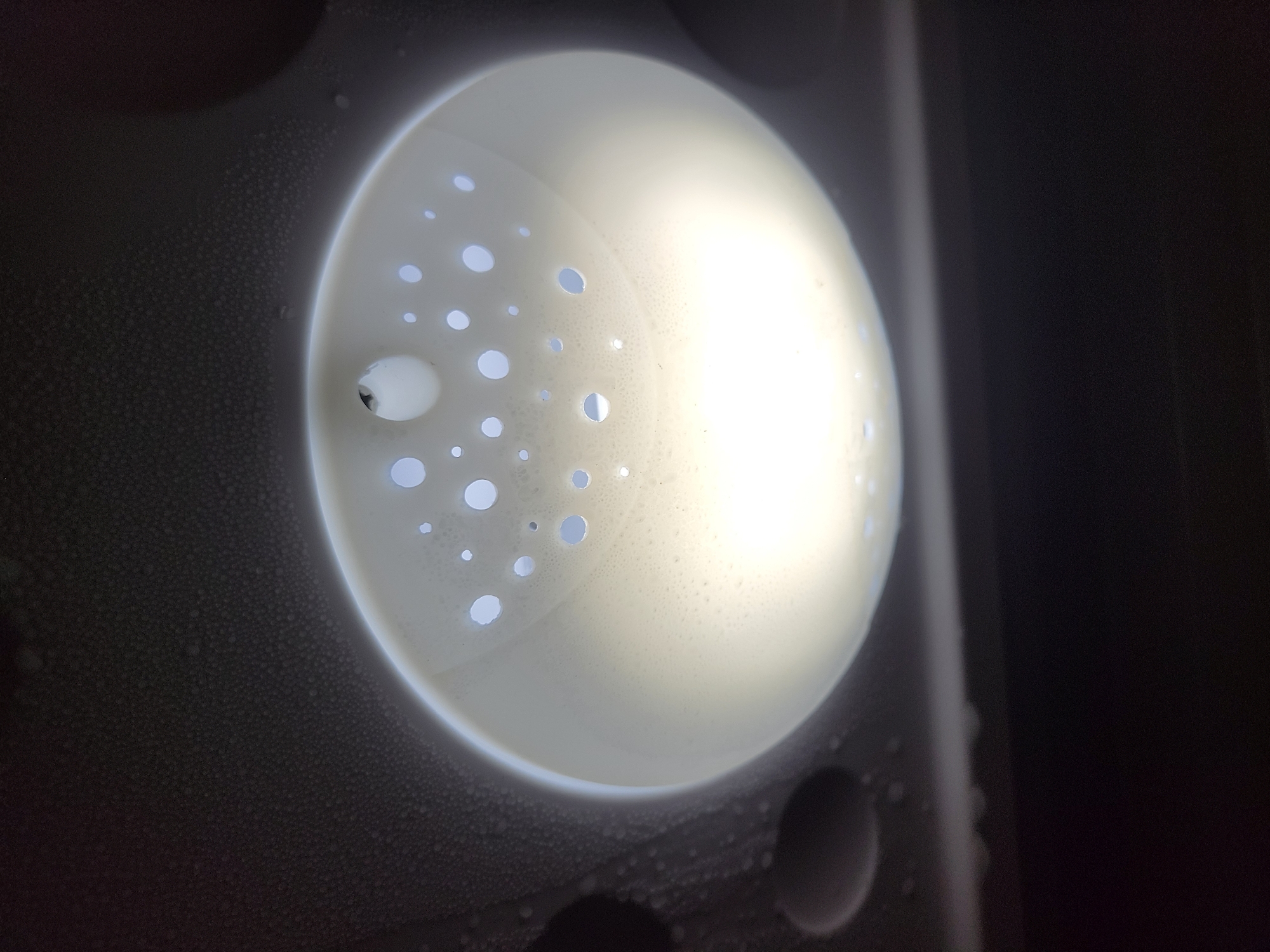 The image shows the inside of a white washing machine drum, with numerous drainage holes. A dark object is attached to the drum wall to the left. Condensation is visible on the dark surfaces surrounding the drum. The location is unknown.