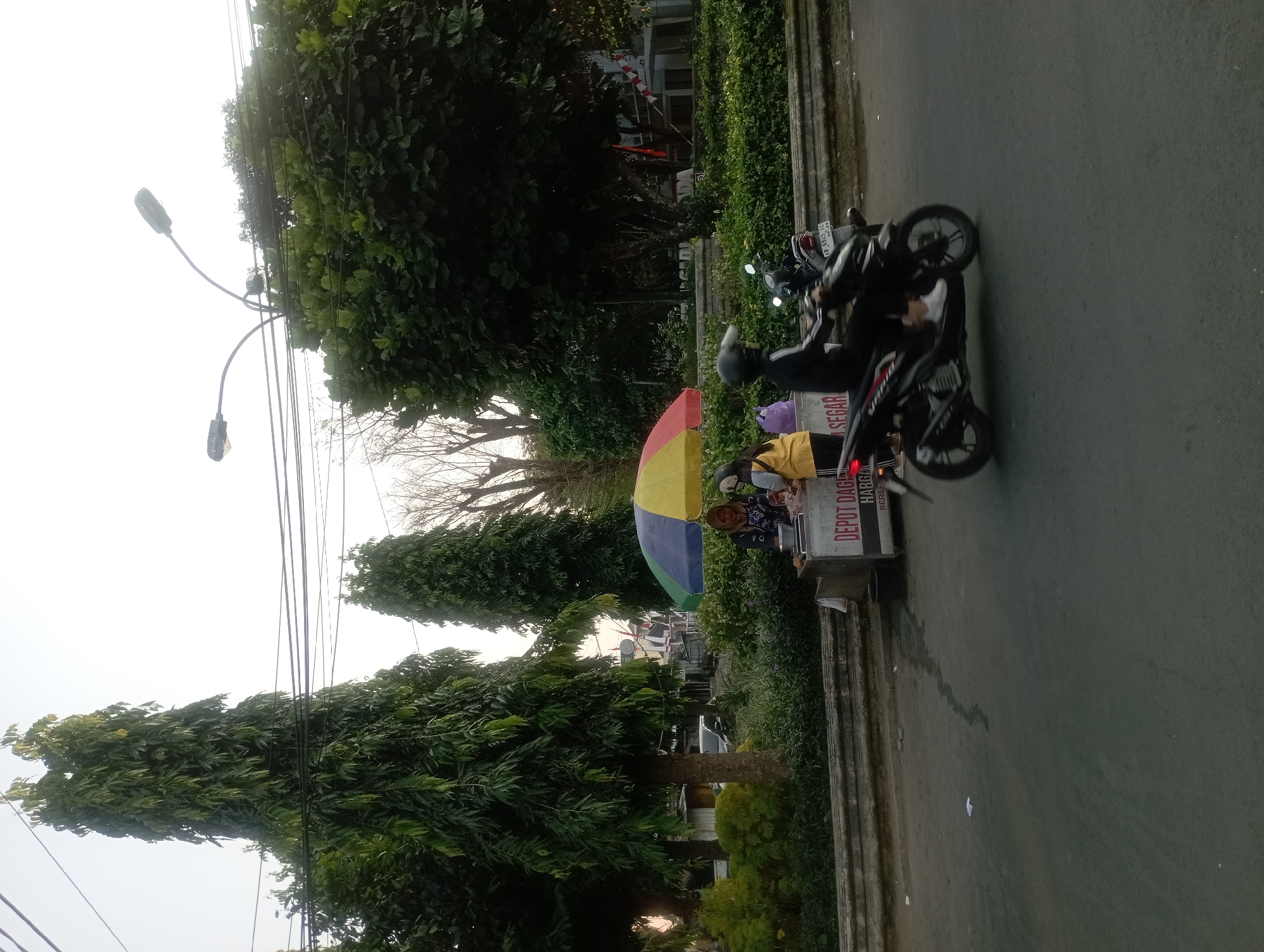 The image shows a street scene in Yogyakarta, Indonesia. A person on a black motorcycle is riding on the right side of the road. In the background, there is a food stall with a colorful umbrella and two people standing behind it. The food stall has a sign that reads "DEPOT DAGE SEGAR HARGA BERSIH". There are trees and streetlights visible in the background, as well as a building. The sky is overcast. The overall impression is a typical street view of a busy city.