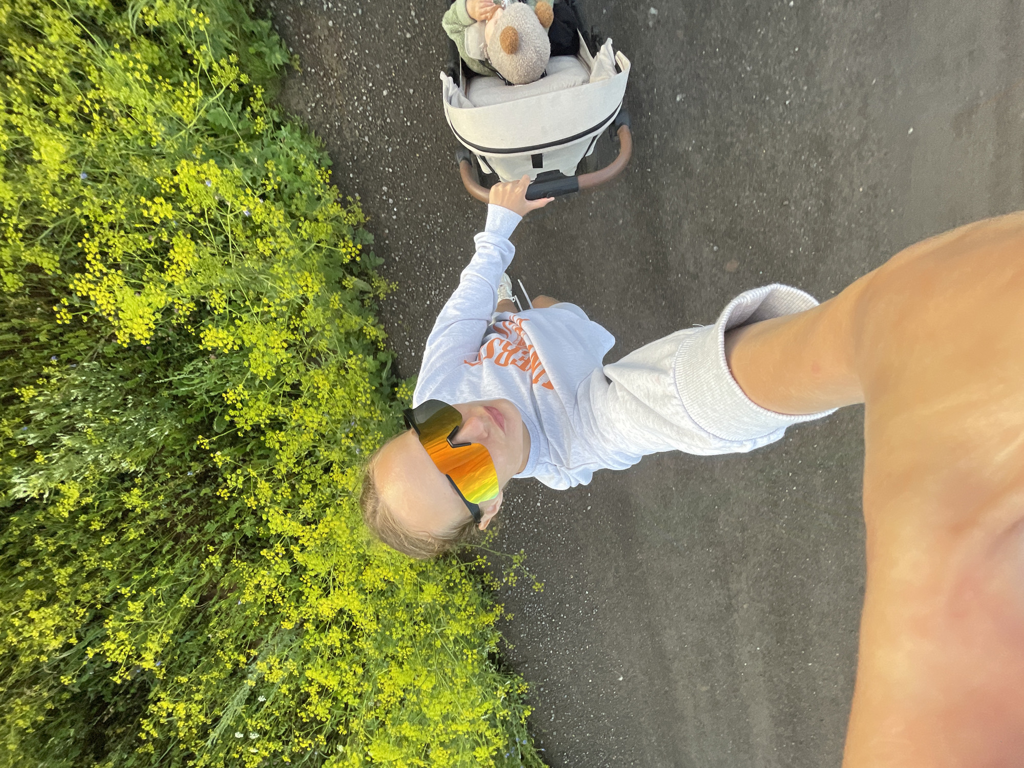 A high-angle, first-person perspective shot shows a person pushing a stroller on a paved path. The person is wearing sunglasses with orange mirrored lenses, a grey sweatshirt with an orange graphic, and white athletic shoes. The stroller is a light beige color with brown handles, and contains a baby wearing a green outfit and a brown teddy bear. To the left of the path, there is dense green foliage with small yellow flowers. The background is an out-of-focus view of similar foliage. The ground appears to be a dark asphalt or paved surface. There are no identifiable landmarks to determine the location in the city of Подшивалово, Russia.