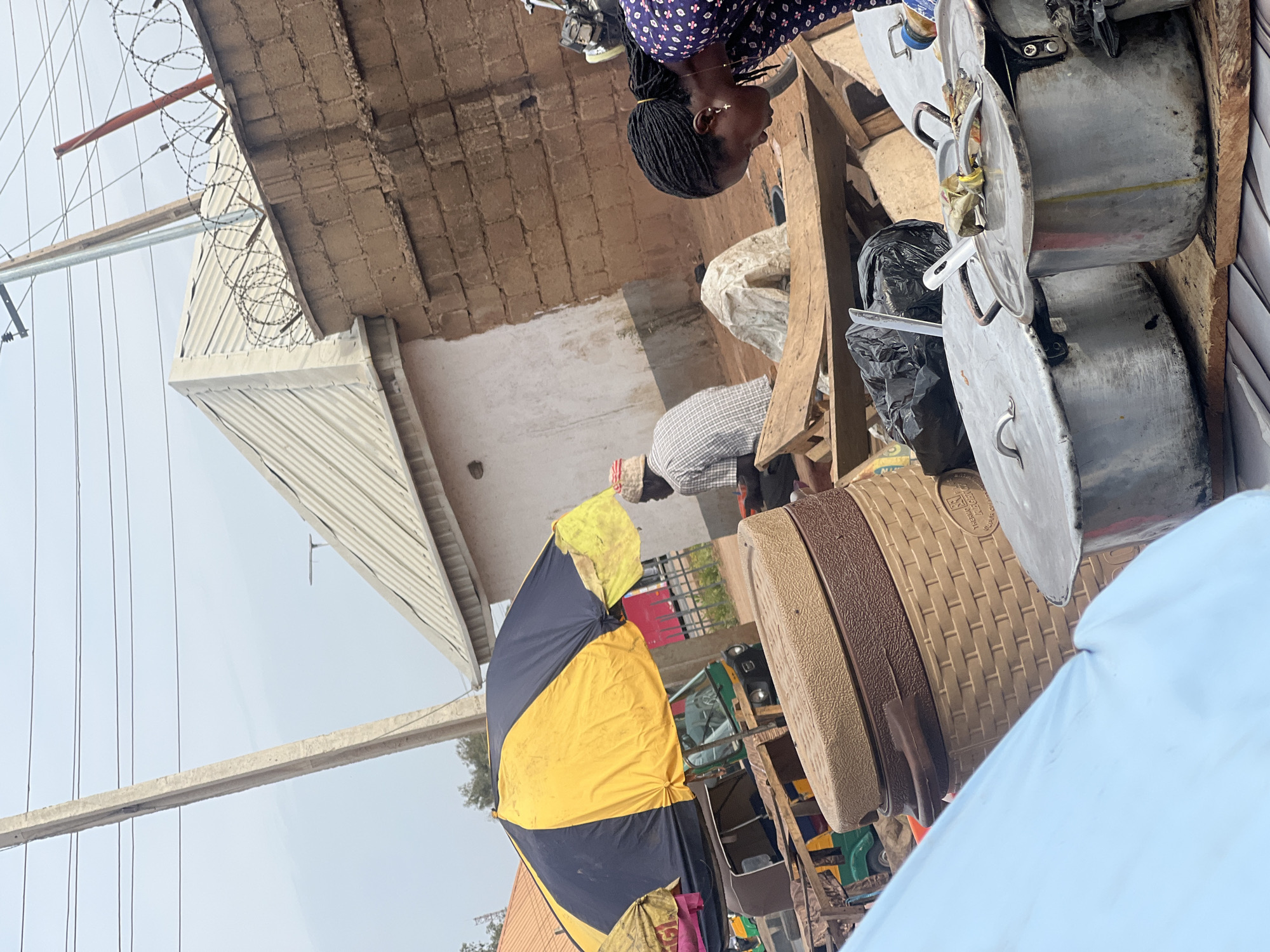 The image depicts an outdoor market scene in Jos, Nigeria, captured during daylight hours under what appears to be an overcast sky.
In the mid-ground, a woman with braided hair, wearing a dark blue patterned top and a gold hoop earring, is visible from the chest up, looking towards the right. Behind her, another individual is seen from the back, wearing a plaid shirt. The setting suggests a bustling atmosphere with goods on display.
To the right, several large, silver-colored aluminum pots with lids are stacked, likely used for cooking or sale. Adjacent to these are woven plastic containers, one of which has the faint text "THERMO BREEZE" visible on its surface. A black plastic bag is also seen among the items. In the lower mid-ground, a large, two-toned umbrella, colored black and yellow, is open, providing shade.
The background features various architectural elements: a building with exposed brickwork and a corrugated metal roof. Coiled barbed wire is prominently visible along the edge of this roof. Overhead, multiple power lines stretch across a light grey sky. Further in the background, a three-wheeled vehicle, resembling a Keke Napep or tricycle taxi, can be glimpsed, indicating a street or thoroughfare nearby. The scene captures daily life, likely a market stall where items are being sold or prepared.