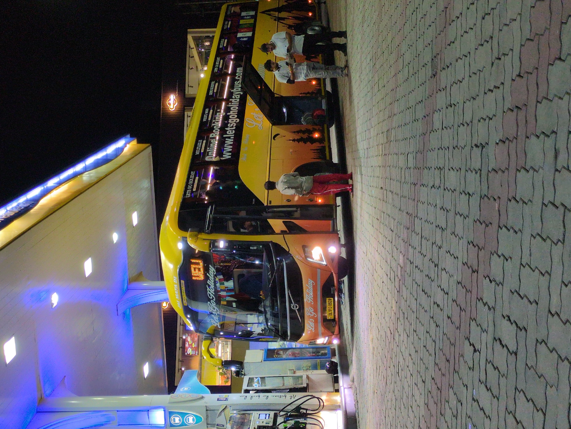 The image captures a brightly lit yellow and orange Volvo bus parked at a fuel station at night. The bus has "Lets Go Holiday" branding and a website address printed on its side. Two men, dressed in casual attire, are standing next to the open storage compartment of the bus, appearing to be engaged in a conversation while looking at their phones. Another man, wearing red pants and a light-colored shirt, stands near the front of the bus. The fuel station features blue and white lighting and modern fuel pumps. The ground is paved with interlocking concrete blocks. In the background, a building with illuminated signage is visible. The scene suggests a temporary stop for the bus, possibly for refueling or passenger pick-up/drop-off. The overall atmosphere is functional and illuminated, typical of a service station operating late at night.