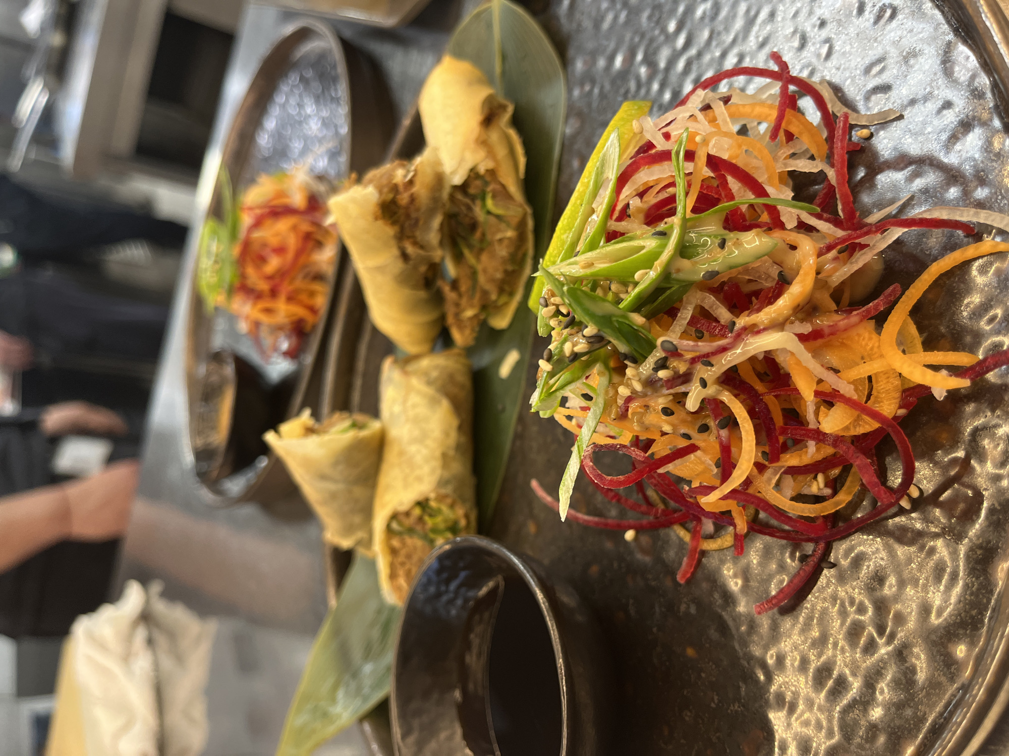 The image displays a close-up, high-angle view of a beautifully plated appetizer or meal, likely in a restaurant setting in Sofia, Bulgaria. The primary subjects are several spring rolls and a vibrant, shredded vegetable salad, presented on dark, textured plates.
On the left, two whole, golden-brown spring rolls are visible, alongside one that has been cut in half, revealing a savory filling composed of dark green and brown elements, suggesting vegetables and possibly meat. These spring rolls are artfully arranged on a large, prominent green leaf, resembling a banana leaf, which adds a natural accent to the presentation. Another spring roll is partially visible further back.
To the right, a colorful mound of finely shredded and spiralized vegetables forms a salad. It features distinct strands of bright red (possibly beets), orange (likely carrots), and white (perhaps daikon radish or white turnip). This salad is garnished with diagonally sliced green onions or chives, a sprinkle of both white and black sesame seeds, and a small wedge of lime. The vegetables appear lightly dressed, giving them a slight sheen.
The plates themselves are dark with a distinctive, hammered or textured surface, which catches the light. In the immediate foreground, a part of a small, dark cup or bowl is visible, likely intended for a dipping sauce. The background is largely out of focus, showing blurred metallic surfaces and indistinct shapes. A person's arm and hand are faintly visible on the far left, and one of the reference crops highlights a gloved hand, suggesting a professional kitchen or serving environment. No text is visible in the image.