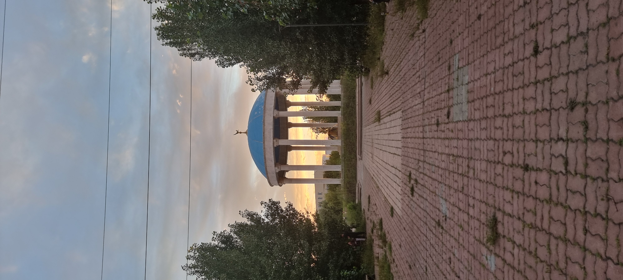 The image depicts a sunset scene at the "Central Park" in Aktobe, Kazakhstan. A blue domed rotunda with white columns stands prominently in the center, topped with a golden bird sculpture. The foreground features a paved walkway made of red interlocking bricks, with patches of grass growing between them. To the left and right of the rotunda are dense green trees. In the background, behind the rotunda, is a white building with the word "ARK" visible on it. The sky is filled with clouds, tinged with orange and yellow hues from the setting sun. Several power lines are visible horizontally across the sky. Two individuals are standing near the left side of the walkway, partially obscured by trees. The overall environment appears to be a public park or plaza.