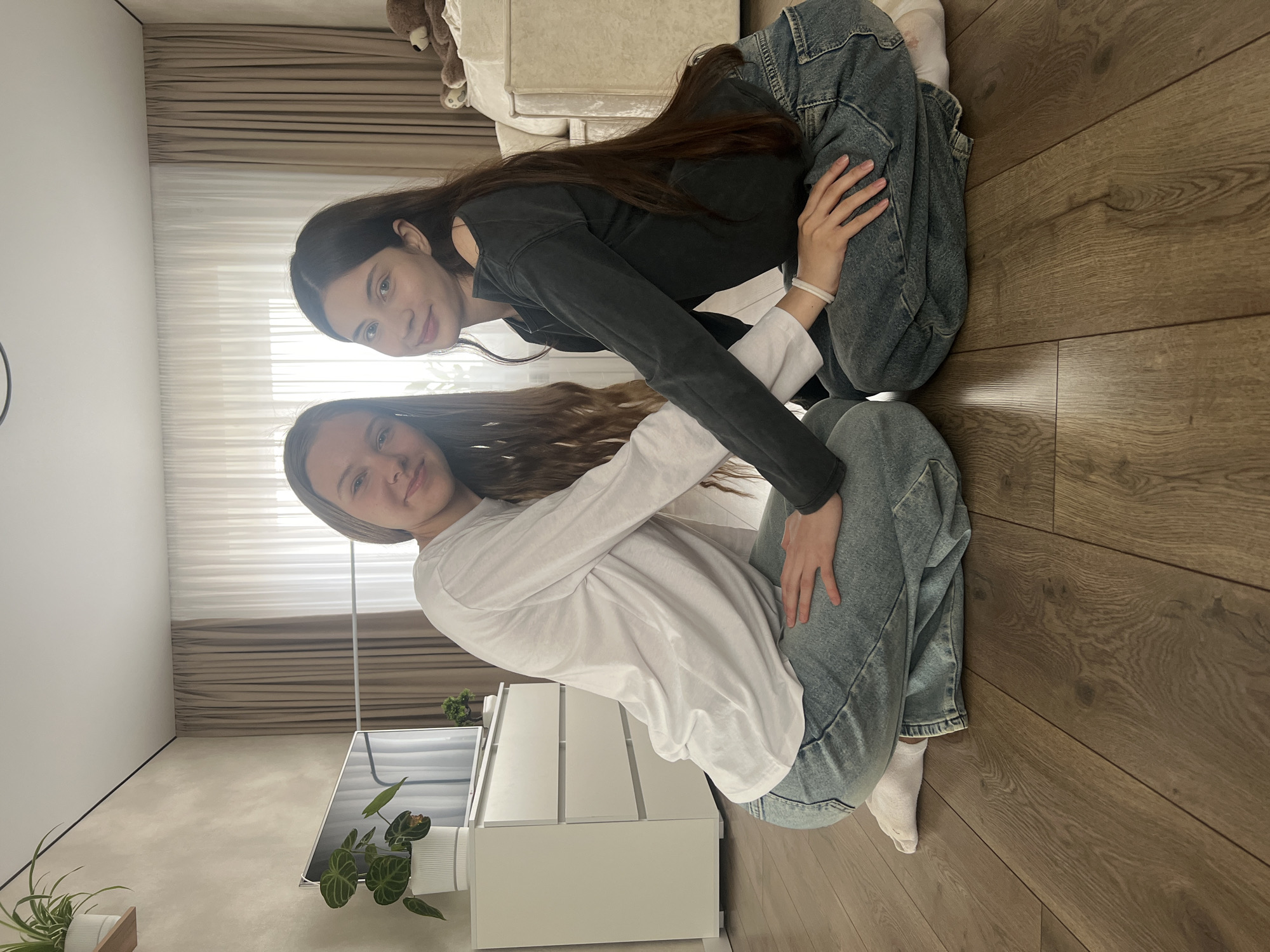 Two young women are crouching on a wooden floor in an indoor setting. One woman wears a white long-sleeved shirt and light-wash jeans, while the other wears a dark long-sleeved shirt and similar light-wash jeans. They are positioned close to each other, with one woman's hand resting on the other's leg. In the background, there is a dresser, a plant, and curtains. A portion of a sofa is visible in the upper background. The location is most likely a residential interior. There is no information to assume the location is in Votkinsk, Russia.