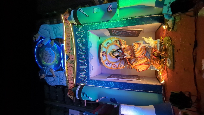 This media file showcases an elaborate, brightly lit festival display, likely a puja pandal, in Dhaka, Bangladesh, at night. The central subject is a richly adorned idol of a Hindu goddess, possibly Saraswati, seated on an ornate, shell-like structure. She is depicted holding a string instrument (likely a veena) and other traditional attributes, surrounded by offerings like flowers and lamps.
The overall theme of the display is a fascinating blend of spirituality and science. On the left, two figures dressed in lab coats, resembling scientists, observe a large, glowing blue sphere containing a brain-like structure illuminated with yellow lights. A prominent, glowing double helix model, representing DNA, runs vertically along the side of the central shrine. Further scientific elements include models of a microscope and a large pen, alongside framed images that appear to be scientific diagrams or texts.
The scene is illuminated by various colored LED lights in blue, green, and yellow, creating a vibrant and celebratory atmosphere against a dark background, indicating nighttime. Visible Bengali text on a vertical banner reads "জ্ঞান ও বিজ্ঞান" (Gyan o Bigyan), translating to "Knowledge and Science," clearly articulating the display's integrated theme. The festive decorations, traditional idol, and modern scientific motifs suggest an event celebrating knowledge in both traditional and contemporary contexts.