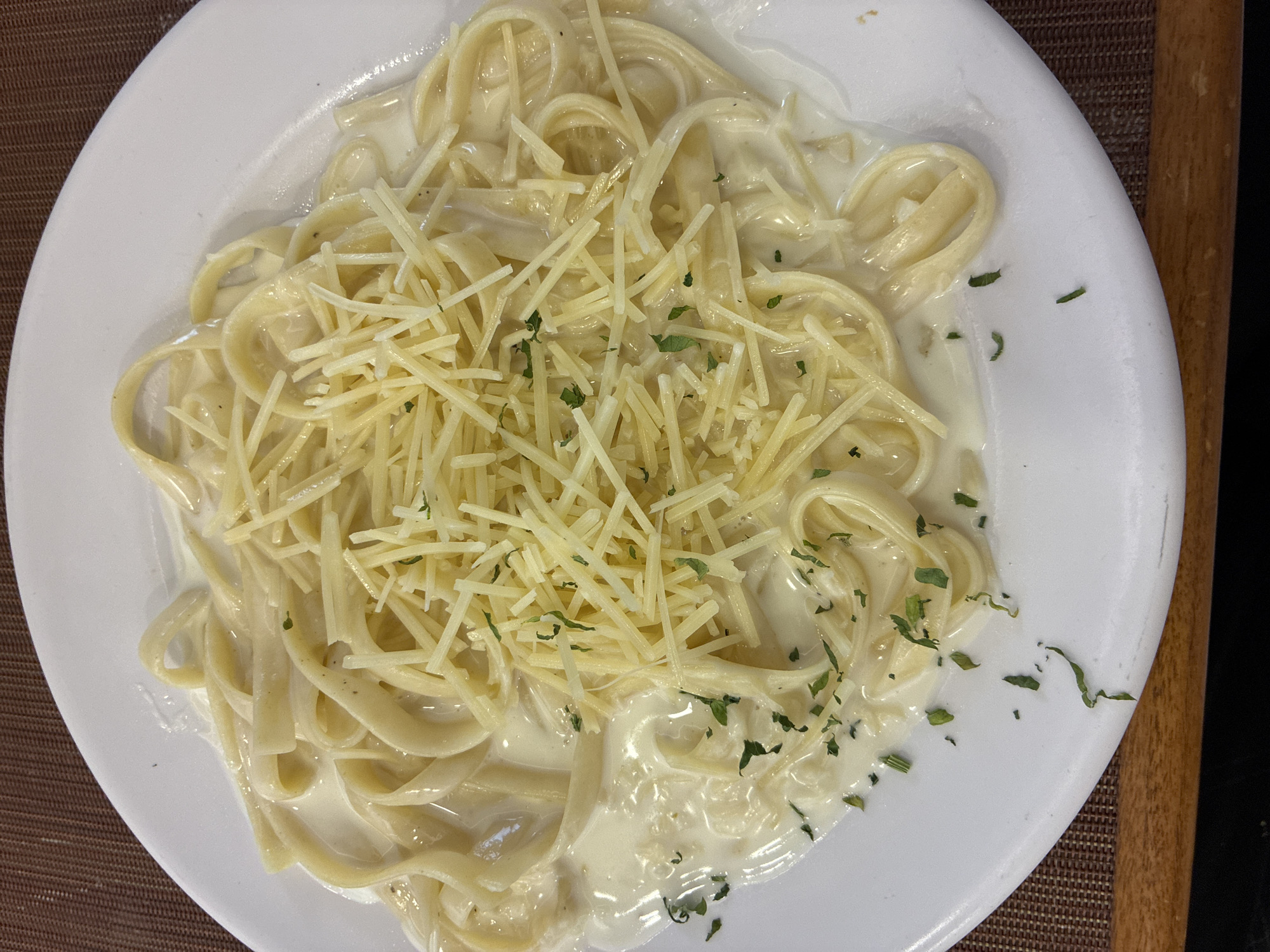 A plate of fettuccine alfredo is presented on a white plate, garnished with shredded cheese and chopped parsley. The plate rests on a surface with a brown and wood pattern. The image was taken in Cuautitlán Izcalli, Mexico.