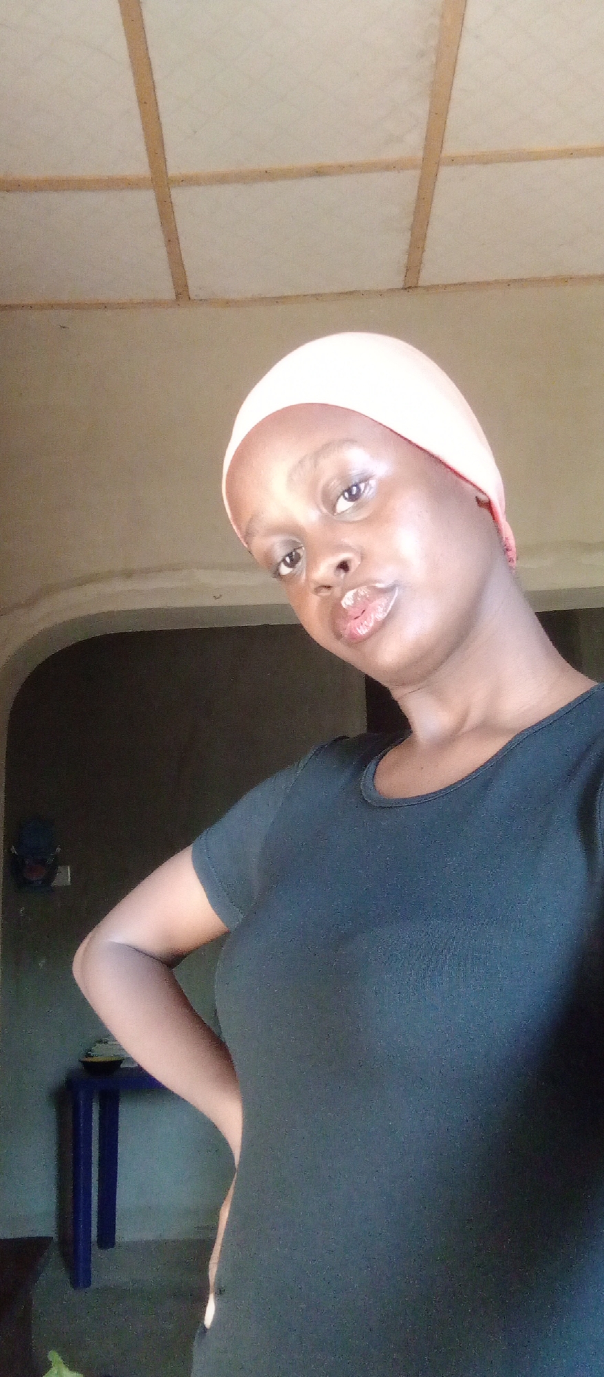 A young woman of African descent is indoors, posing for a selfie. She is wearing a dark-colored shirt and a light pink head covering. Her left hand is on her hip. Behind her is an arched doorway leading into a dimly lit room. A small blue table is visible in the background room. The ceiling is a grid of white panels with wooden support beams. The image was taken in Akwanga, Nigeria.