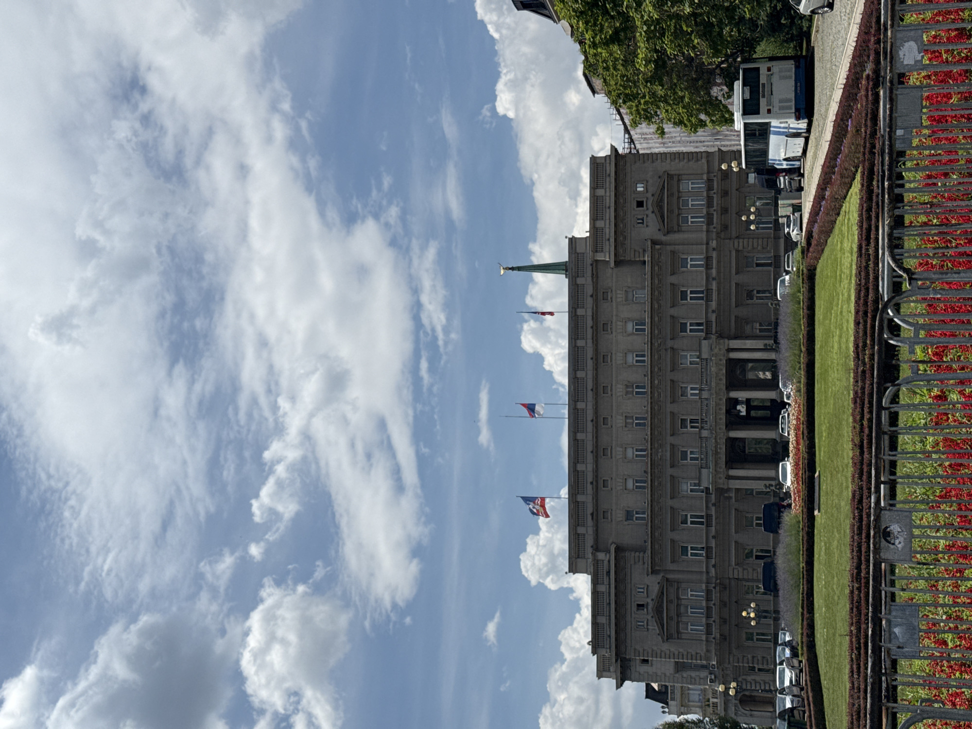 This is a ground-level, eye-level, medium-shot photograph of the Presidency of Serbia building in Belgrade, Serbia, taken on a day with a partly cloudy sky. The building, a large, neoclassical structure with a grey facade, occupies the majority of the frame. It features multiple rows of windows, columns at the entrance, and flags flying from the roof.
In front of the building, a manicured lawn is bordered by flowerbeds and hedges. A metal fence runs across the foreground, separating the viewer from the garden. Several cars and a white bus with blue stripes are parked on the street to the right of the building.
The sky above is filled with white cumulus clouds against a bright blue backdrop. The overall scene depicts a public space in a European city, likely a government or administrative center, on a bright, clear day.