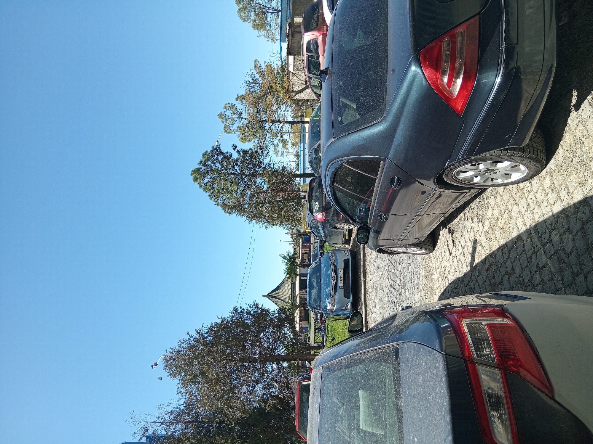 The image shows a row of parked cars on a cobblestone street, under a clear blue sky. The cars are of various makes and colors, including a dark blue Mercedes and a grey car with red taillights. Trees are visible in the background, and beyond them, there is a glimpse of a building with a distinct oriental-style roof, along with other structures. The location is likely a parking area near a public space or building in Batumi, Georgia. No people are visible.