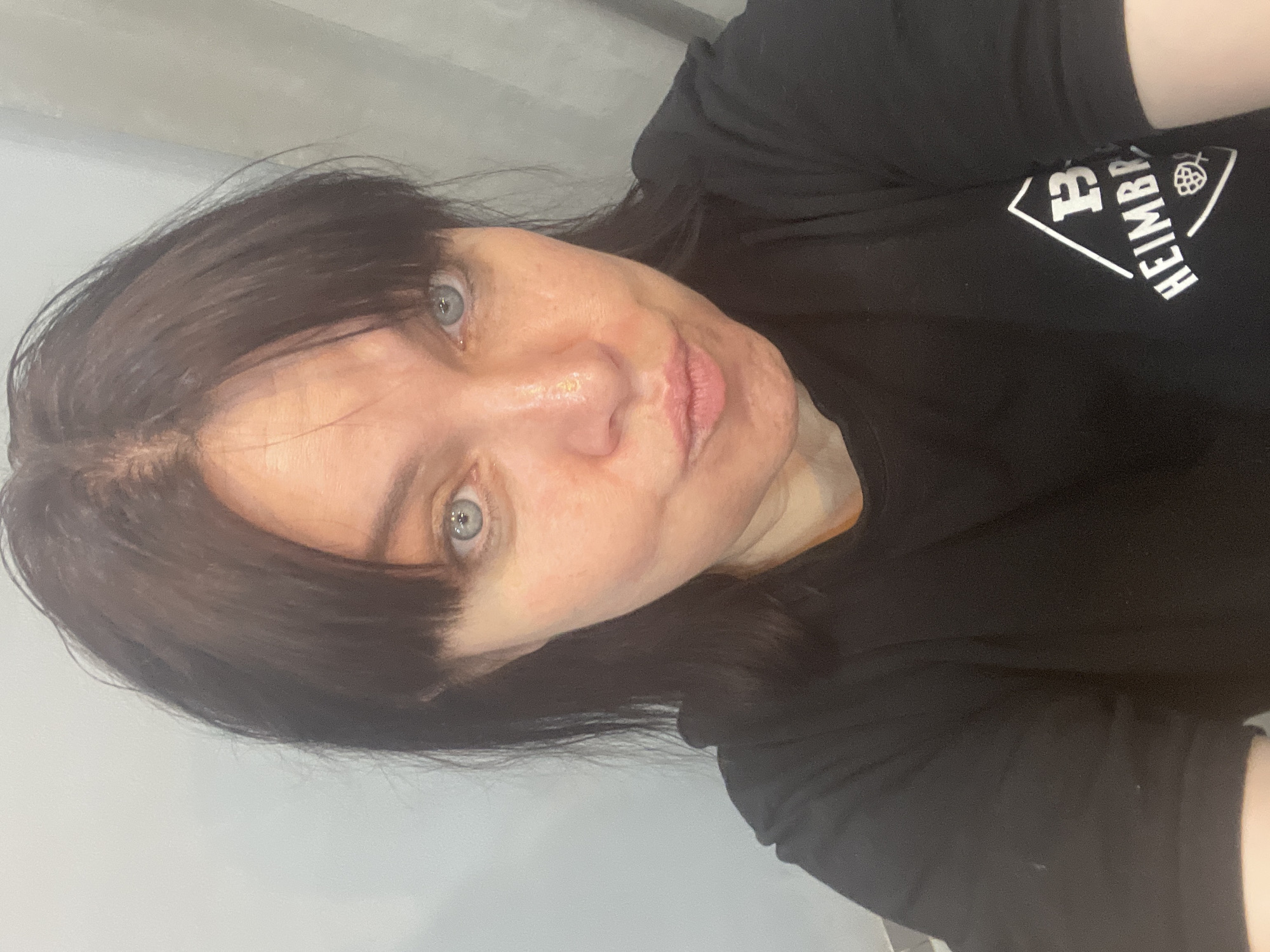A medium-shot, front-facing selfie of a woman with fair skin, dark brown hair, and blue eyes. She has bangs and her hair is pulled back. She is wearing a black t-shirt with a white logo on the right side of the chest. The logo is a diamond shape with the letters "B" and "H" above the word "HEIMBR," and a hop cone below. The background is a light gray wall and a curtain. The image appears to have been taken indoors. The woman's lips are pursed.