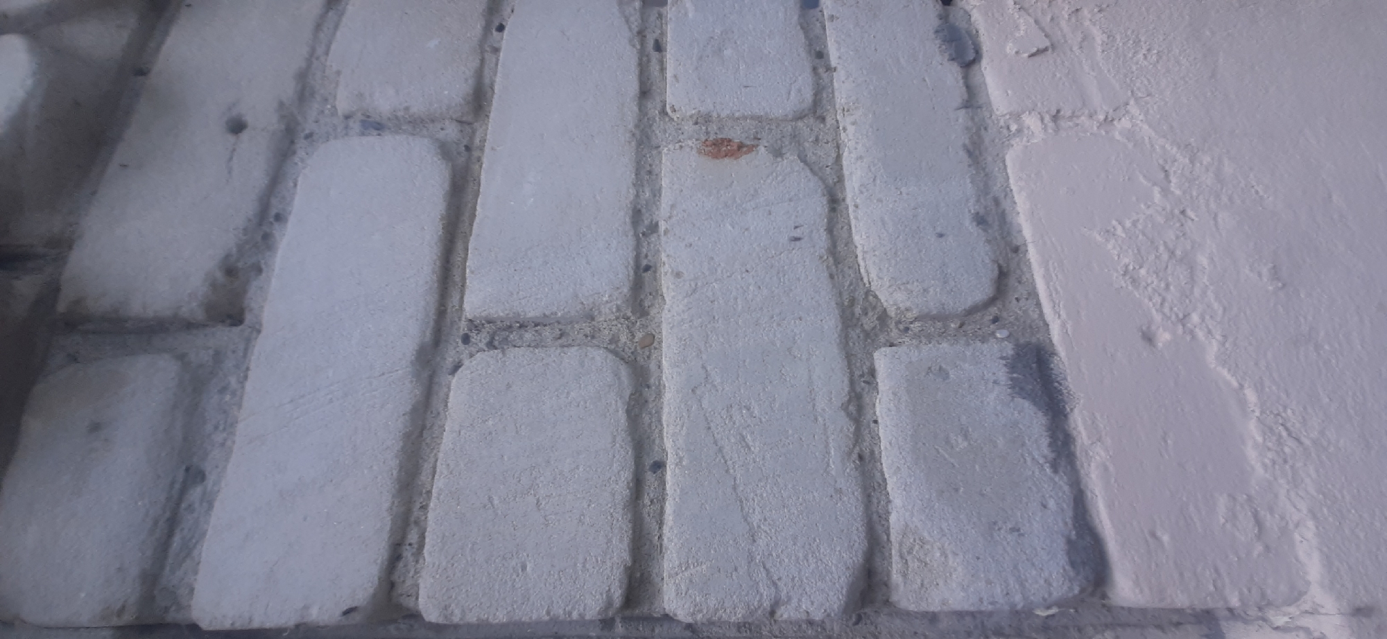 The image shows a close-up, angled view of a painted brick wall. The bricks are a light gray color, and the mortar between them is a darker gray. Some of the bricks have slight imperfections and variations in texture. The bottom portion of the image shows a band of a lighter, possibly pinkish-white paint applied over the bricks. There are no people or discernible actions taking place. The scene appears to be an exterior wall, possibly of a building. The location in Yessentukskaya, Russia, is not visually indicated in the image itself.