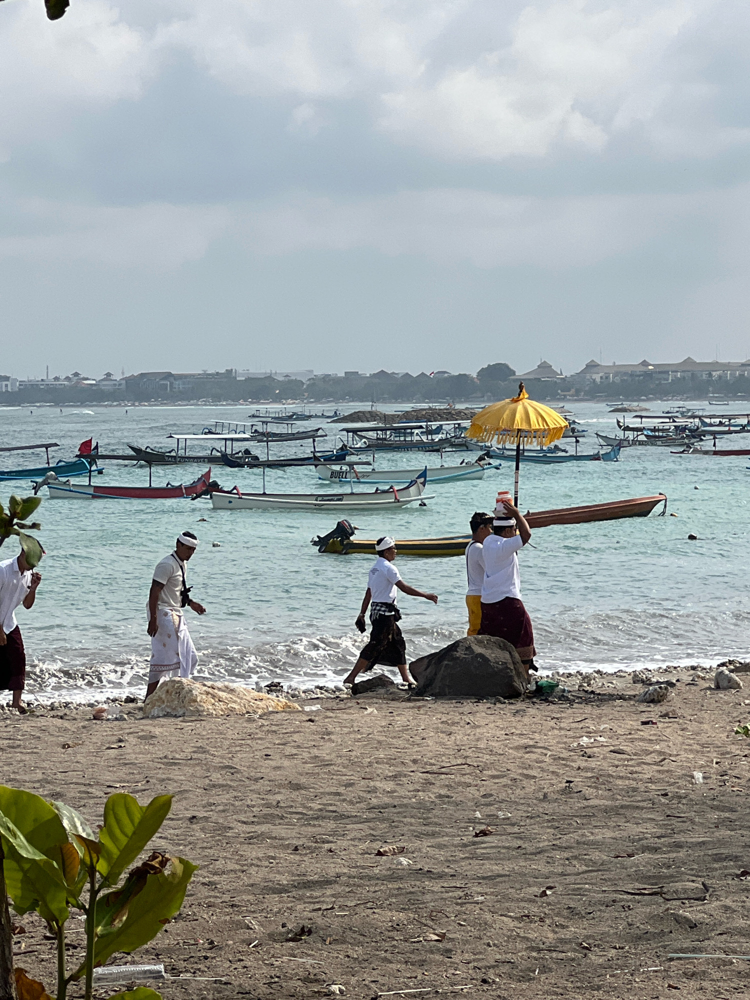A group of men in traditional Balinese attire are walking along the shore of Kuta Beach in Kuta, Indonesia. The men are dressed in white shirts and sarongs, and some are wearing white headbands. One man is carrying a yellow umbrella, which is open and providing shade. Behind the men, numerous traditional Balinese fishing boats are anchored in the turquoise waters. The beach is sandy, with some scattered rocks and debris. In the background, a town with buildings and structures is visible along the coastline. The sky is overcast with gray clouds.