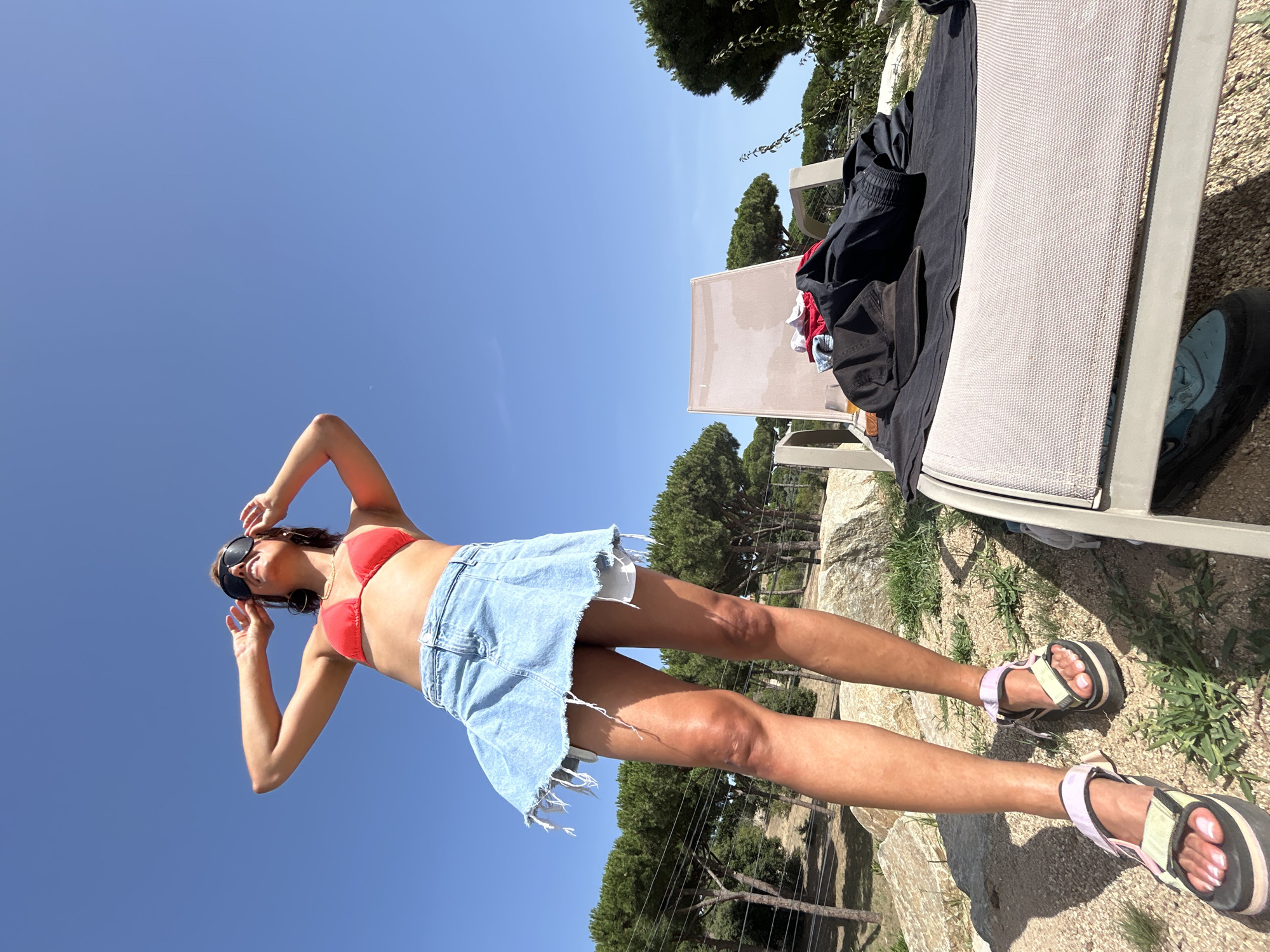 A low-angle, full-body shot captures a woman standing on a rocky, outdoor terrain with a clear blue sky above. She is wearing a red bikini top, a distressed denim mini-skirt, and platform sandals. Her hands are positioned near her sunglasses, and she is looking upwards. In the background, a lounge chair is laden with folded clothing, including a black cap and what appears to be a dark jacket or pants, next to a glass. Pine trees and some fencing are visible further in the background. The setting is likely a private residence or a resort area near Sant Vicenç de Montalt, Spain, characterized by dry, rocky ground and Mediterranean vegetation. The lighting suggests a sunny day, casting shadows on the ground.