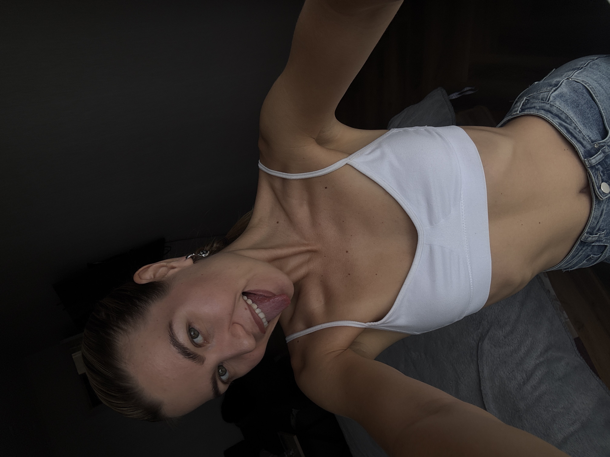 A woman with fair skin is wearing a white top and blue jeans, and sticking her tongue out. She is indoors, in a room with a dark wall. The location is Unknown, Unknown.