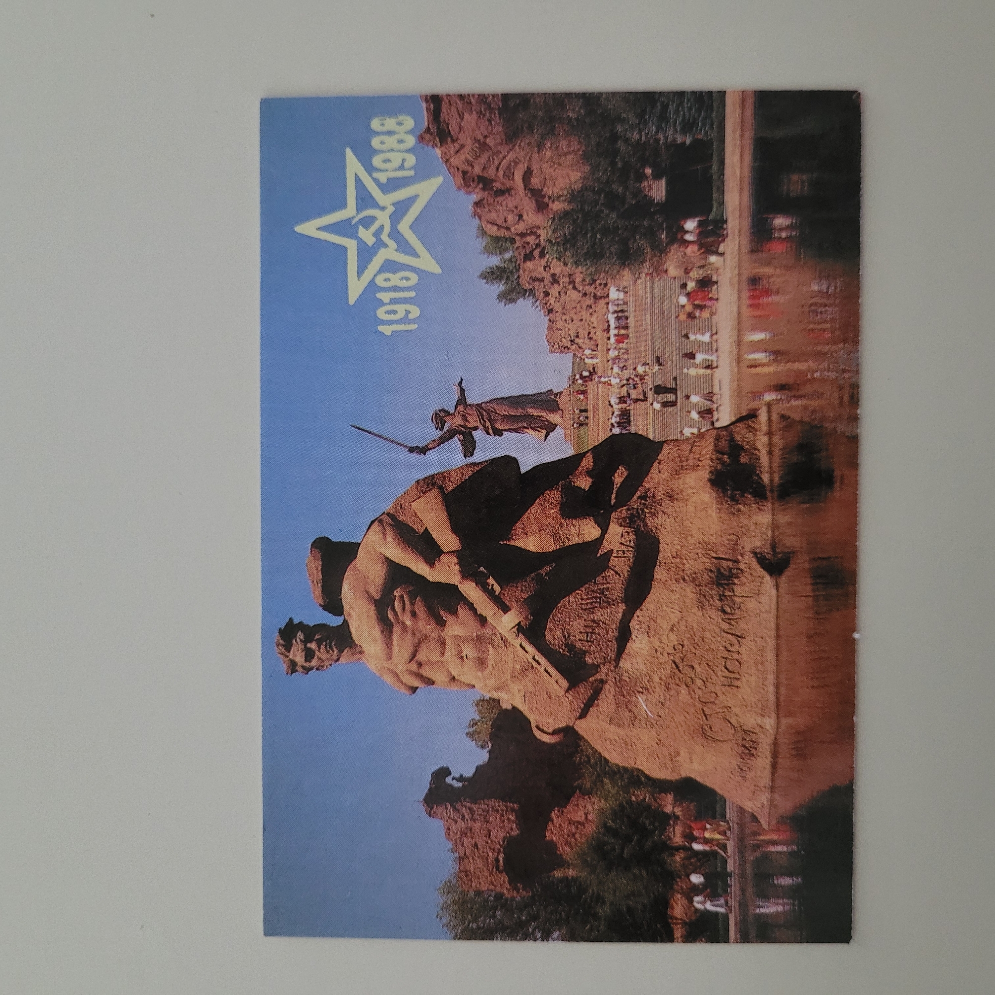The image is a postcard featuring a monument of two soldiers. The soldier in the foreground is muscular and holds a rifle. Behind him, another soldier is partially visible. The monument is situated in front of a large staircase leading up to a statue of a woman holding a sword. There are people in the background, some walking on the stairs and others gathered at the bottom. The monument is reflected in the water in front of it. To the right of the monument, a star with a hammer and sickle inside is visible, along with the years "1918" and "1988". The postcard itself has a white border and a light blue background in the top right corner. This monument is the "Motherland Calls" statue, located in Volgograd, Russia, not Jūrmala, Latvia.