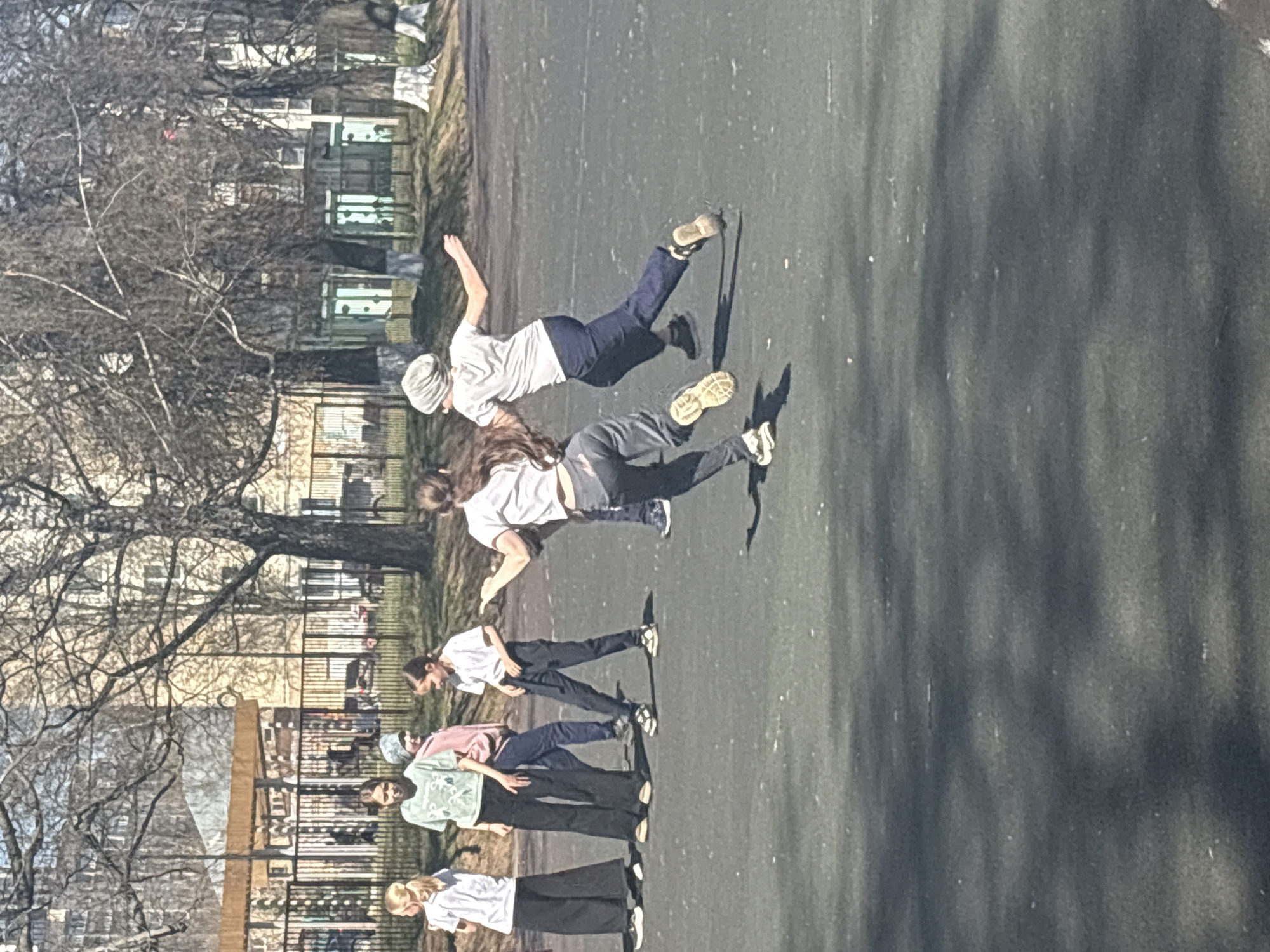 The image depicts a group of people in what appears to be an urban outdoor setting. Six individuals are standing or walking on an asphalt surface, while two are in mid-air, seemingly jumping or dancing. All individuals are wearing casual clothing, including t-shirts and pants. The setting includes trees, a fence, and buildings in the background, suggestive of a park or public space. Based on the provided information that the image is located in Izhevsk, Russia, the buildings' architecture is consistent with that geographic location.