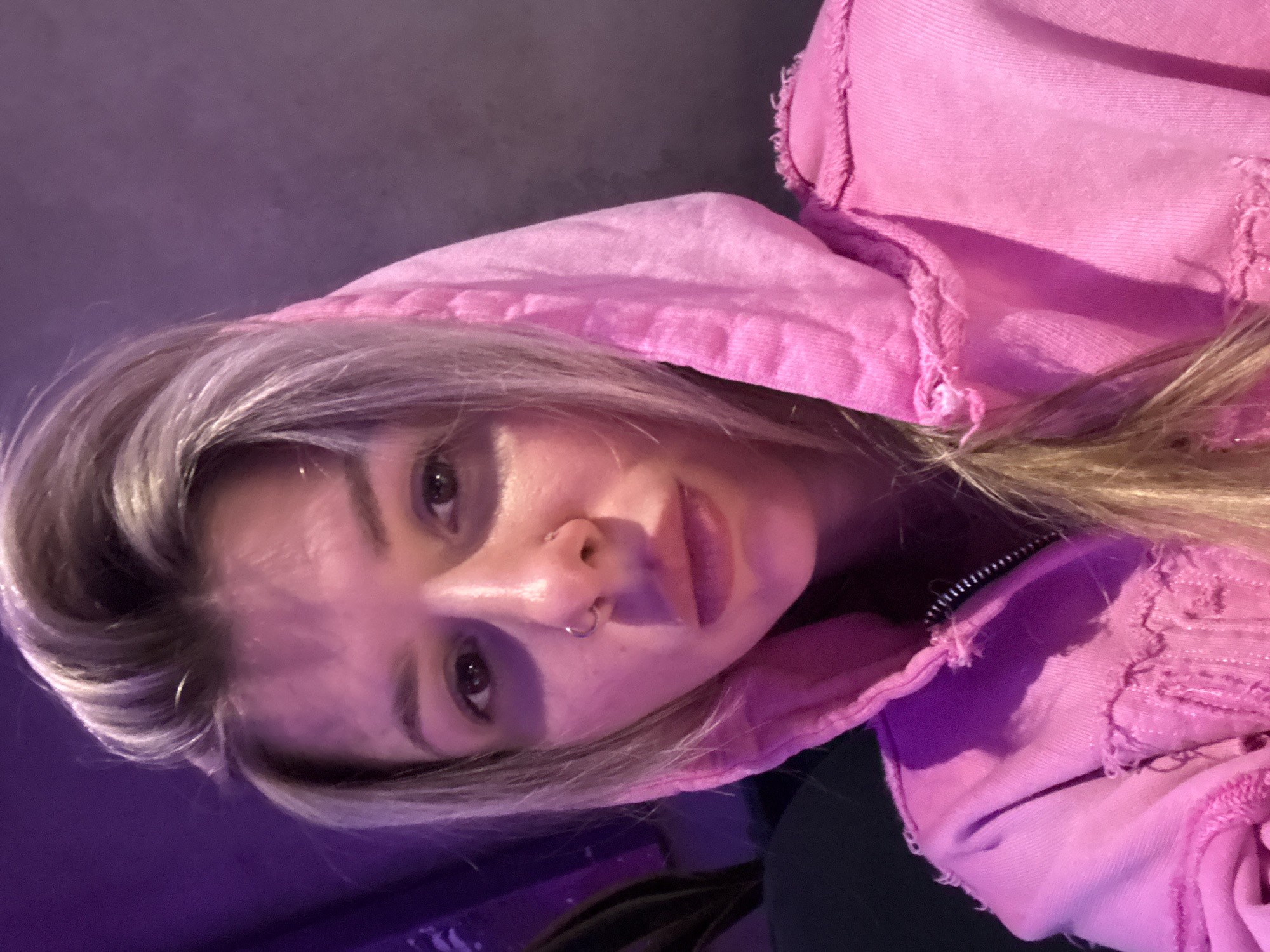 A close-up, medium shot captures a young woman with blonde hair pulled back. She is wearing a pink hooded sweatshirt with a zipper. The lighting is purple and pink, casting shadows on her face. She has a nose ring and appears to be looking directly at the camera. The background is dark and out of focus. The scene is indoors.