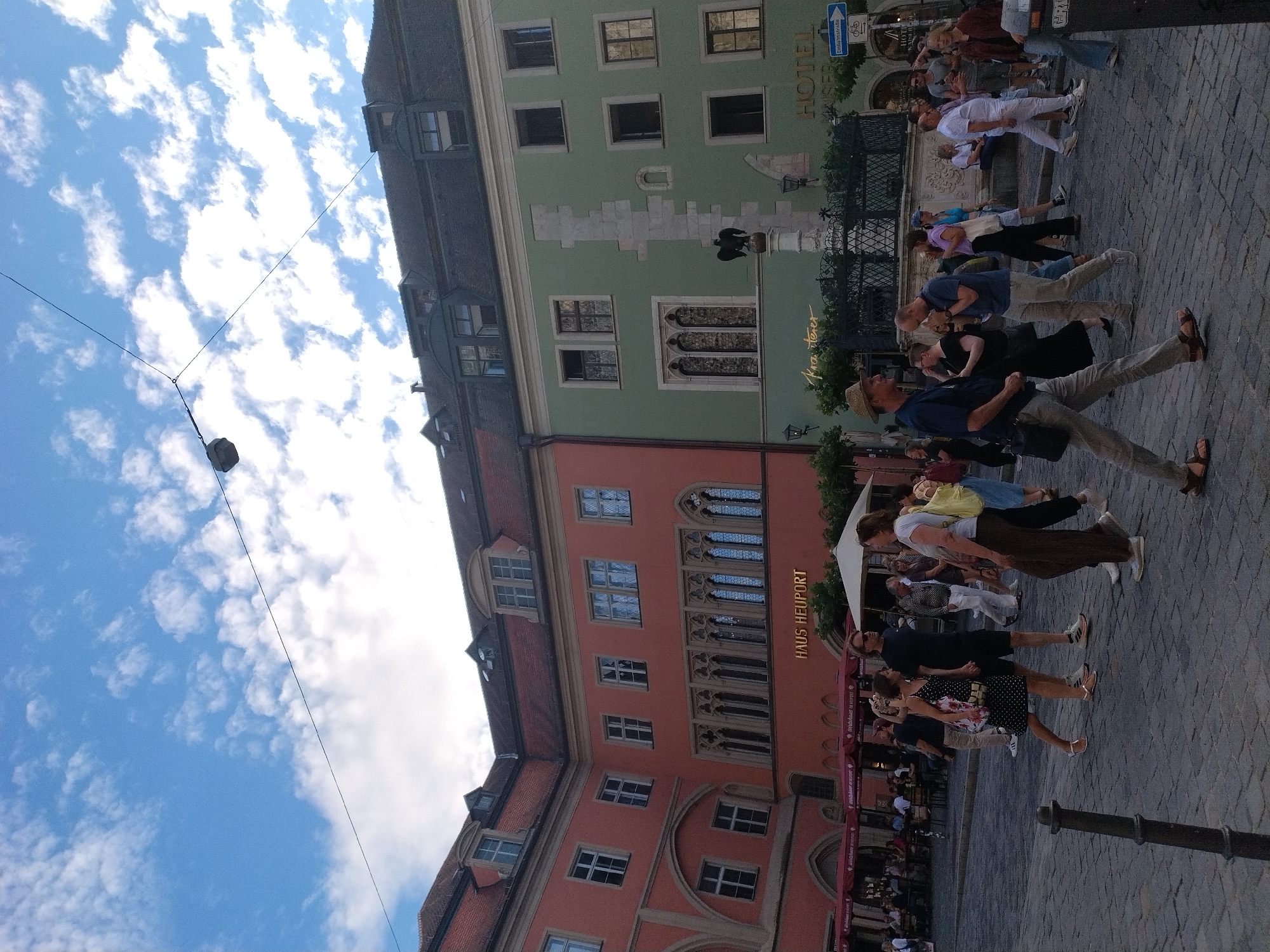 The image depicts a European city square with cobblestone paving. In the background, two buildings with distinct colors stand side-by-side: a salmon-pink building on the left and a light green building on the right. Both buildings exhibit traditional European architecture with multiple stories, windows, and dormers. Signs on the buildings read "HAUS HEUPORT" and "HOTEL KAISER..." respectively. The sky above is a bright blue with scattered white clouds.
In the foreground, a group of people are walking in various directions, suggesting a public space. The people are dressed in casual summer attire. Some individuals are walking together, while others appear to be walking alone or in small groups. A lamppost and a street sign are also visible in the scene. The overall impression is of a bustling urban environment, likely a popular tourist or public gathering area.
Specifically, this scene captures the **Haidplatz** in Regensburg, Germany. The distinctive pink and green buildings are characteristic landmarks of this historic square.