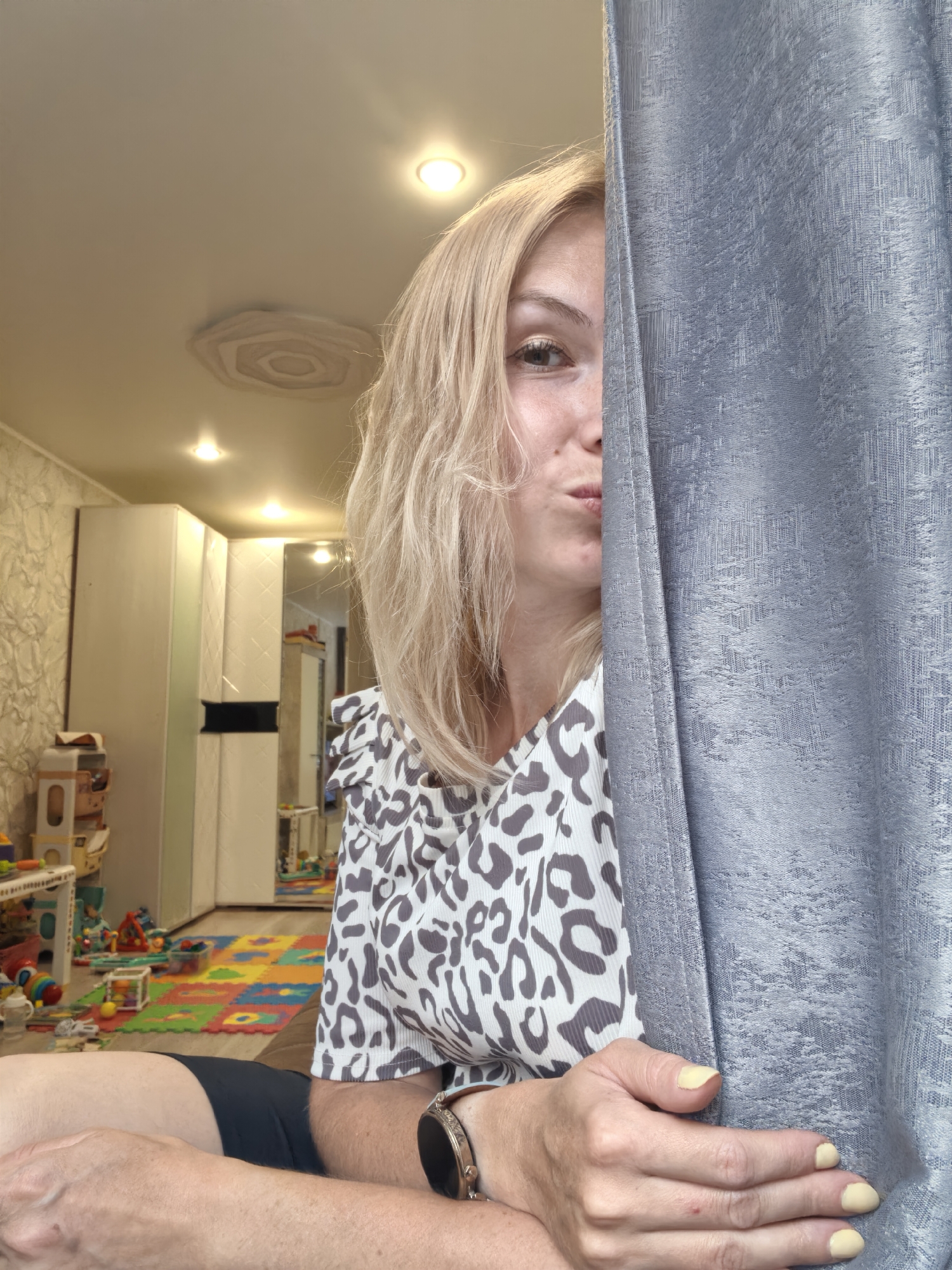 The image is a medium shot of a woman peeking from behind a gray curtain, with her face turned towards the camera. She has blonde hair and is wearing a white top with a leopard print pattern. Her right hand, with light yellow nail polish, rests on the curtain. A smartwatch is visible on her left wrist.
The background suggests an indoor setting, likely a child's room, given the presence of a colorful puzzle mat on the floor and various toys scattered around. There's a white wardrobe and a shelf unit with more toys visible. The ceiling has recessed lighting and an unusual decorative element.
The lighting appears to be artificial, indicating it could be daytime or evening. The woman's expression is playful, with her lips pursed. The overall impression is a casual, candid moment captured at home in Sterlitamak, Russia.